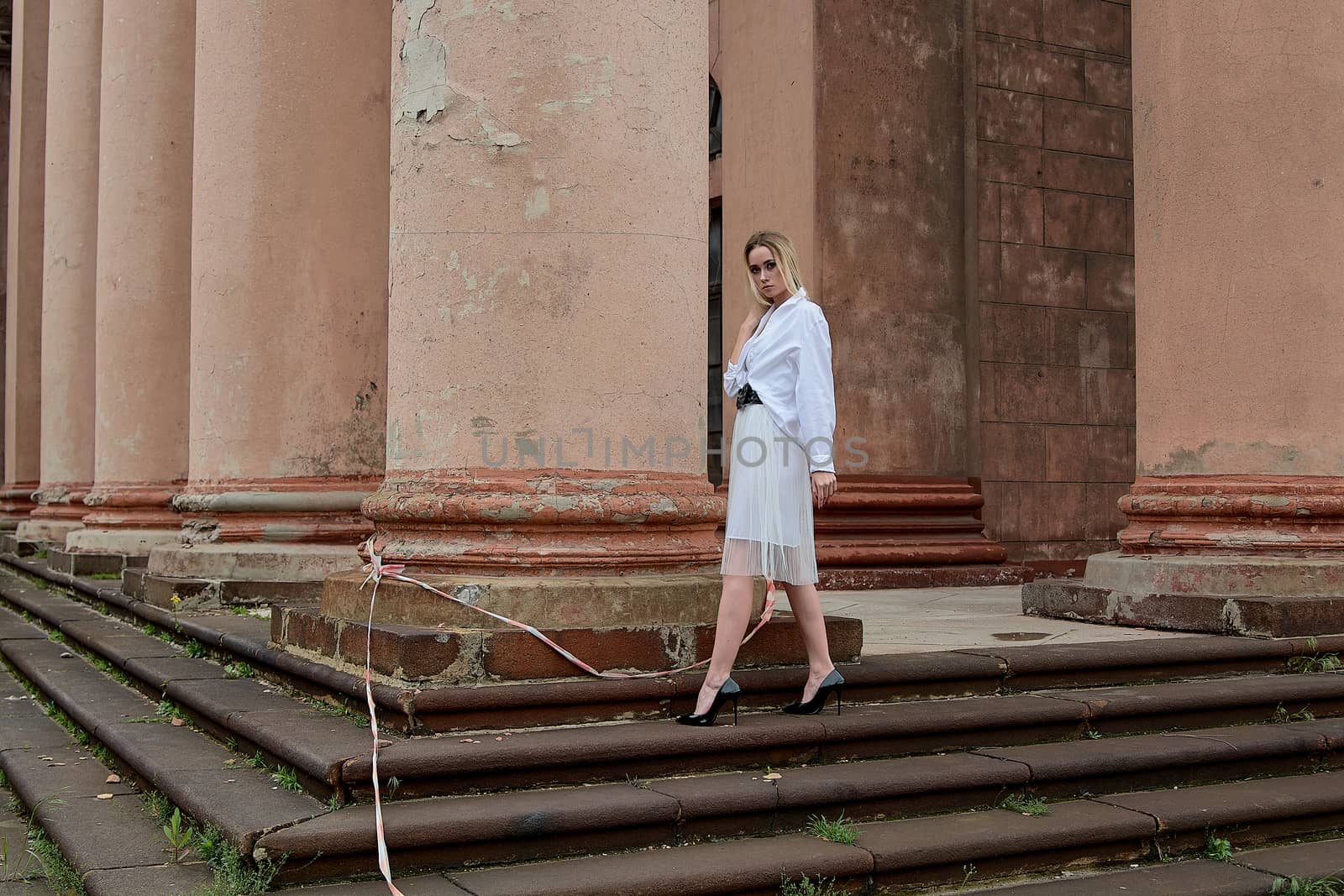 Young blonde woman dreses in white skirt and shirt posing on the steps of the old looking vintage building with large 
columns and bas-reliefs. Fashion look's woman. Young woman's modern portrait.