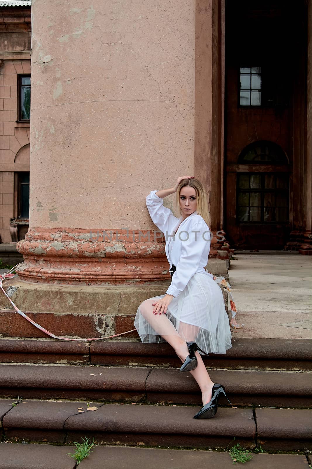 Young blonde woman dreses in white skirt and shirt posing on the steps of the old looking vintage building with large 
columns and bas-reliefs. Fashion look's woman. Young woman's modern portrait.