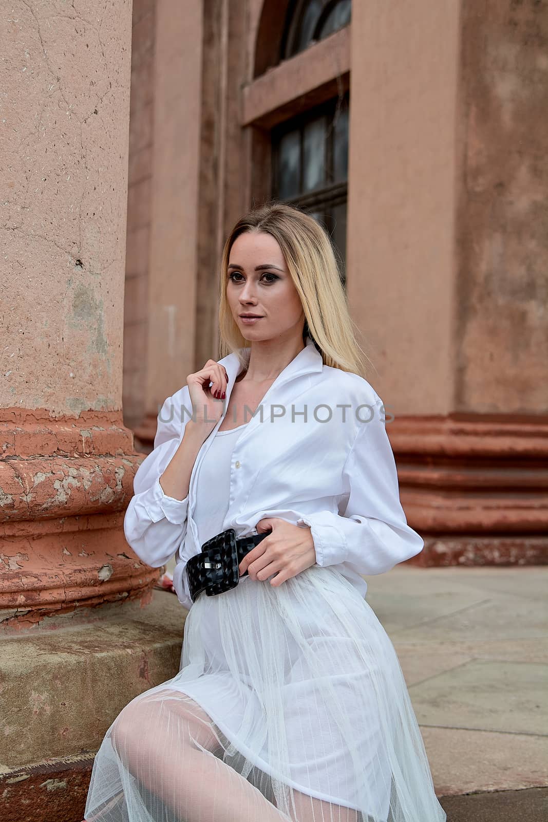 Young blonde woman dreses in white skirt and shirt posing on the steps of the old looking vintage building with large 
columns and bas-reliefs. Fashion look's woman. Young woman's modern portrait.