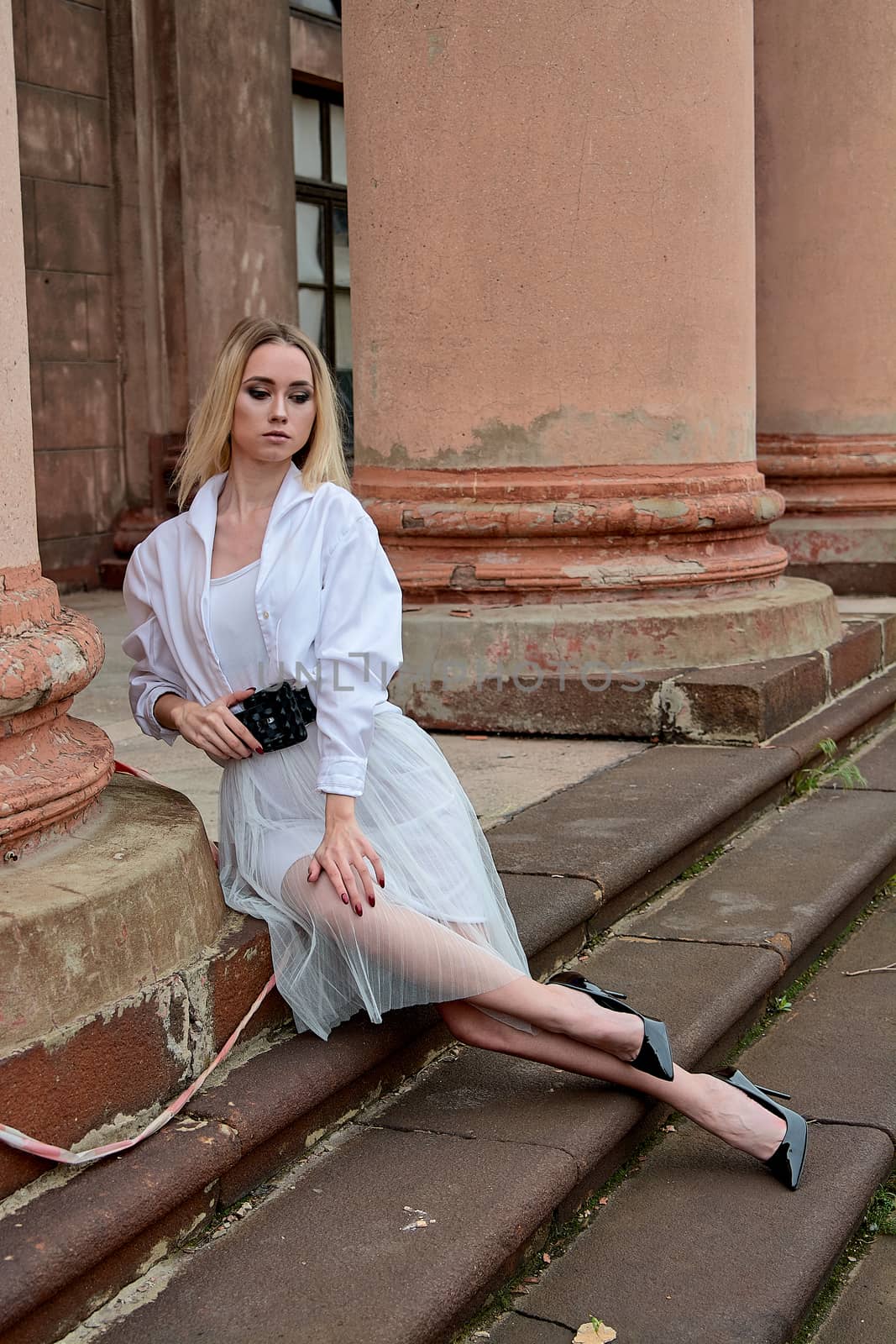 Young blonde woman dreses in white skirt and shirt posing on the steps of the old looking vintage building with large 
columns and bas-reliefs. Fashion look's woman. Young woman's modern portrait.