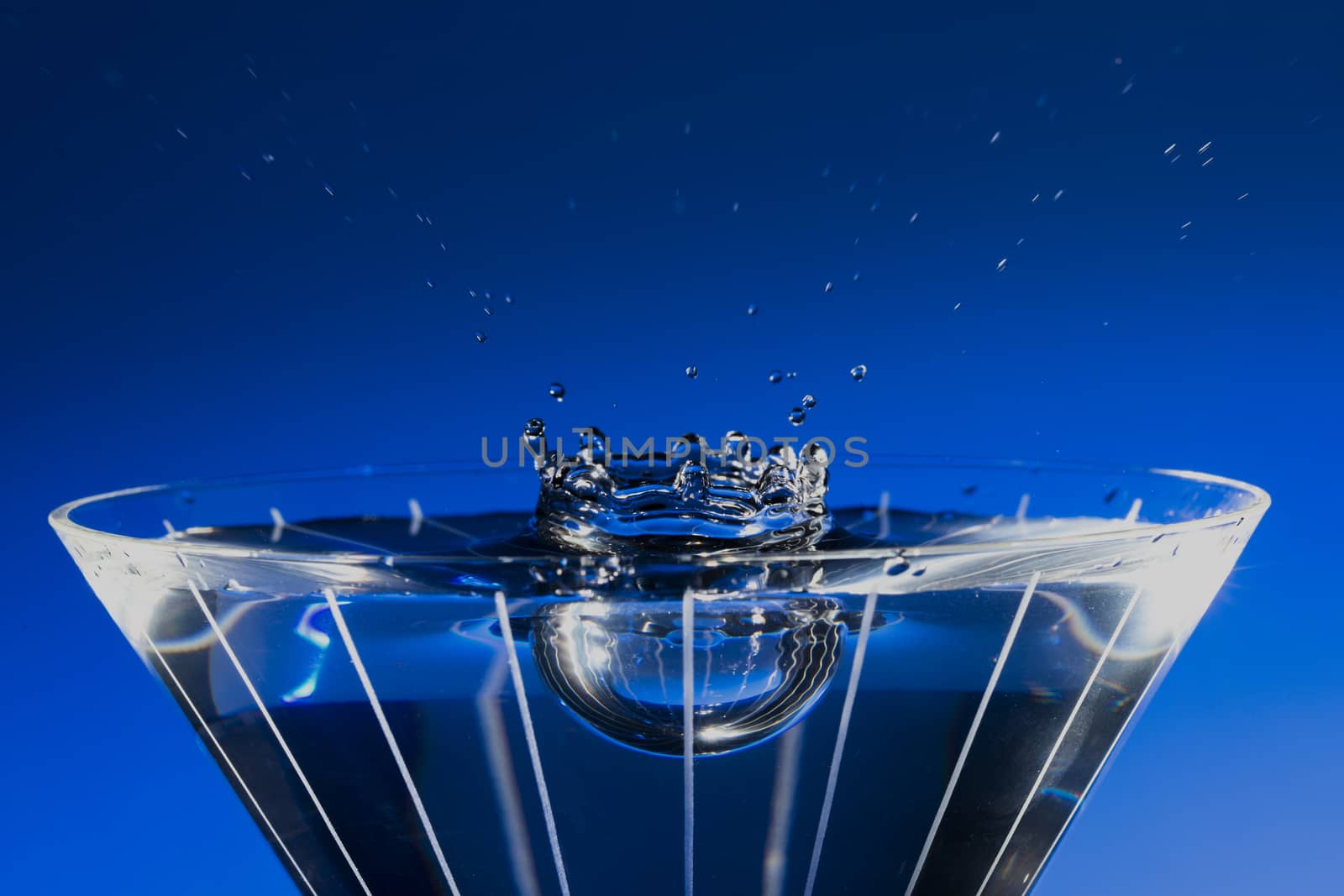 Stop motion of water drop splashing down into filled martini glass, with blue background.