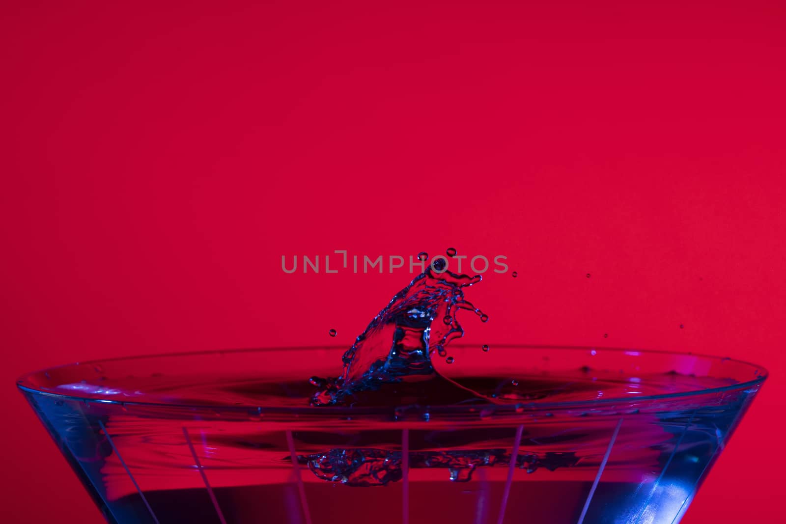 Droplets collide over a martini glass with red background and blue liquid highlights.
