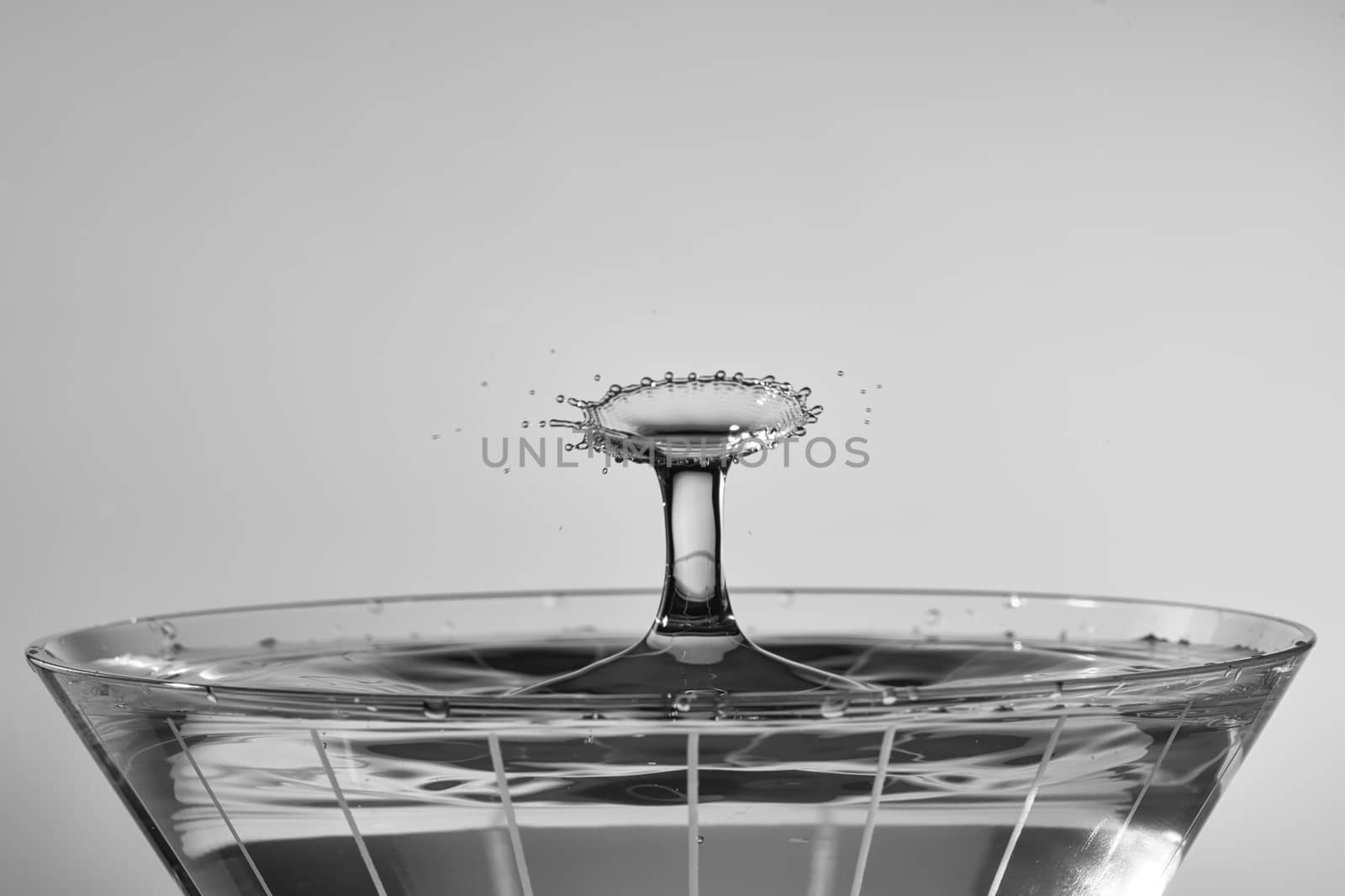 Droplets collide above martini glass, creating a radial fan pattern. Processed as monochrome.
