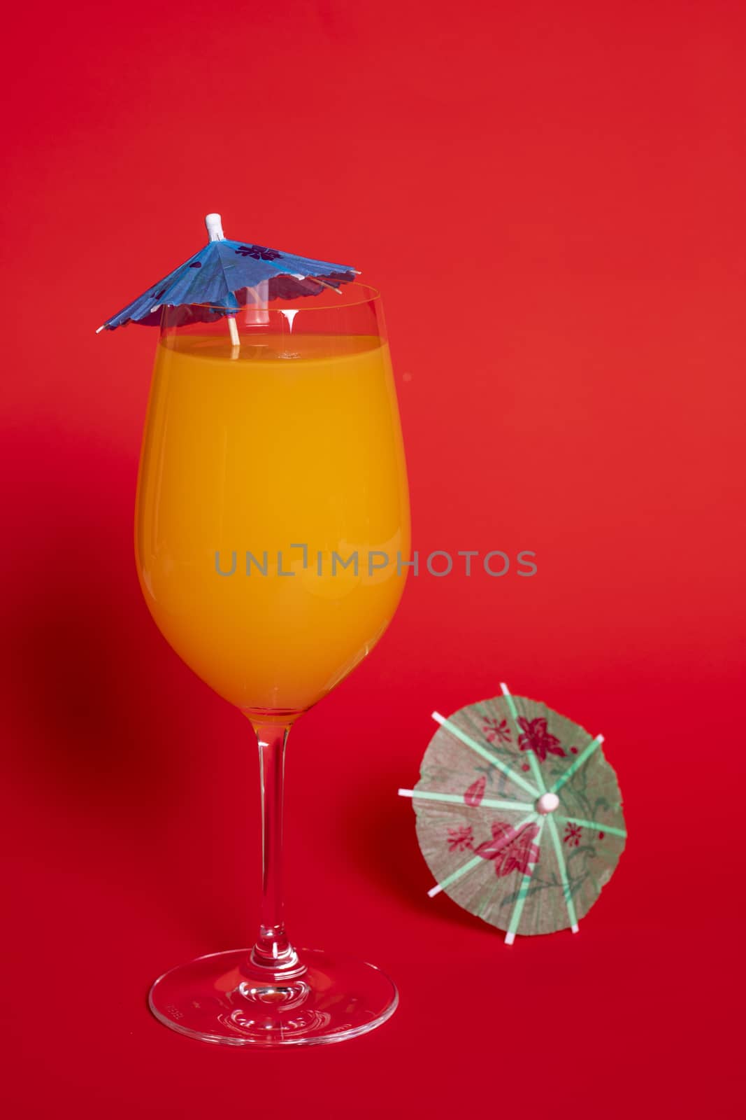 Orange drink with a blue umbrella in a wine glass set against a solid red background. A green umbrella lies beside the glass.