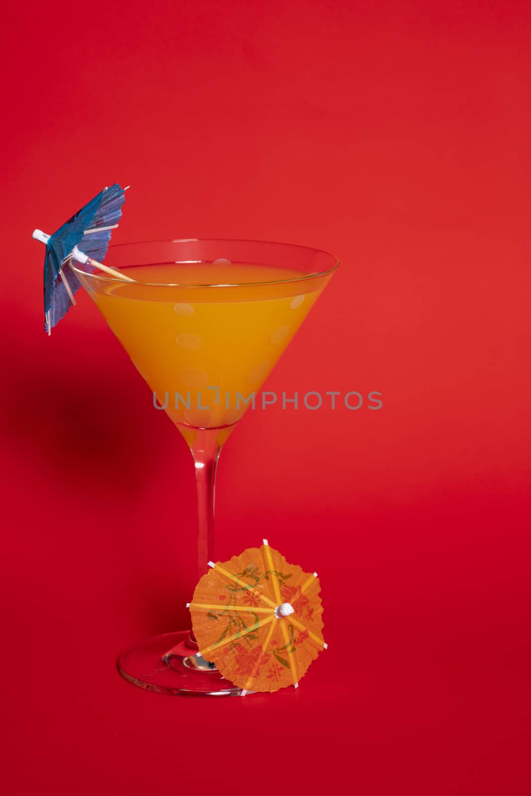 Orange drink with a blue umbrella in a martini glass set against a solid red background. An orange umbrella lies at the base of the glass.