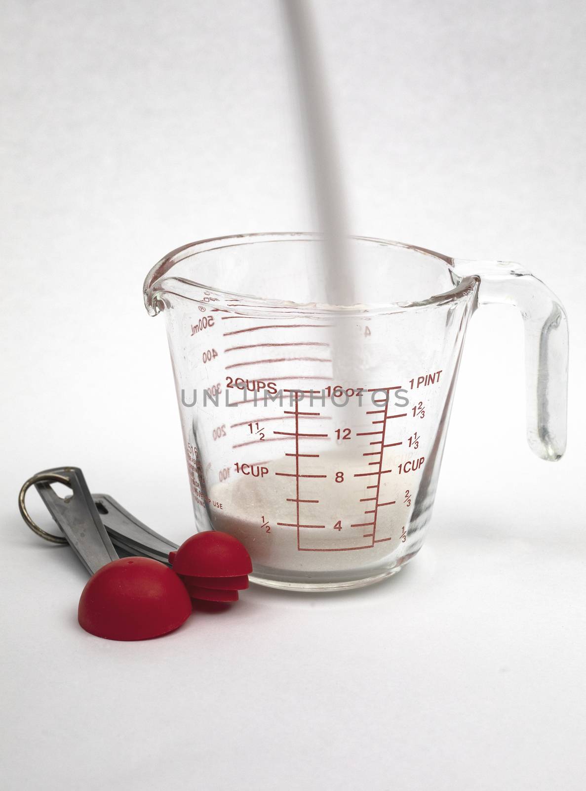 Pouring white cane sugar into a measuring cup with measuring spoons in view, time exposure.