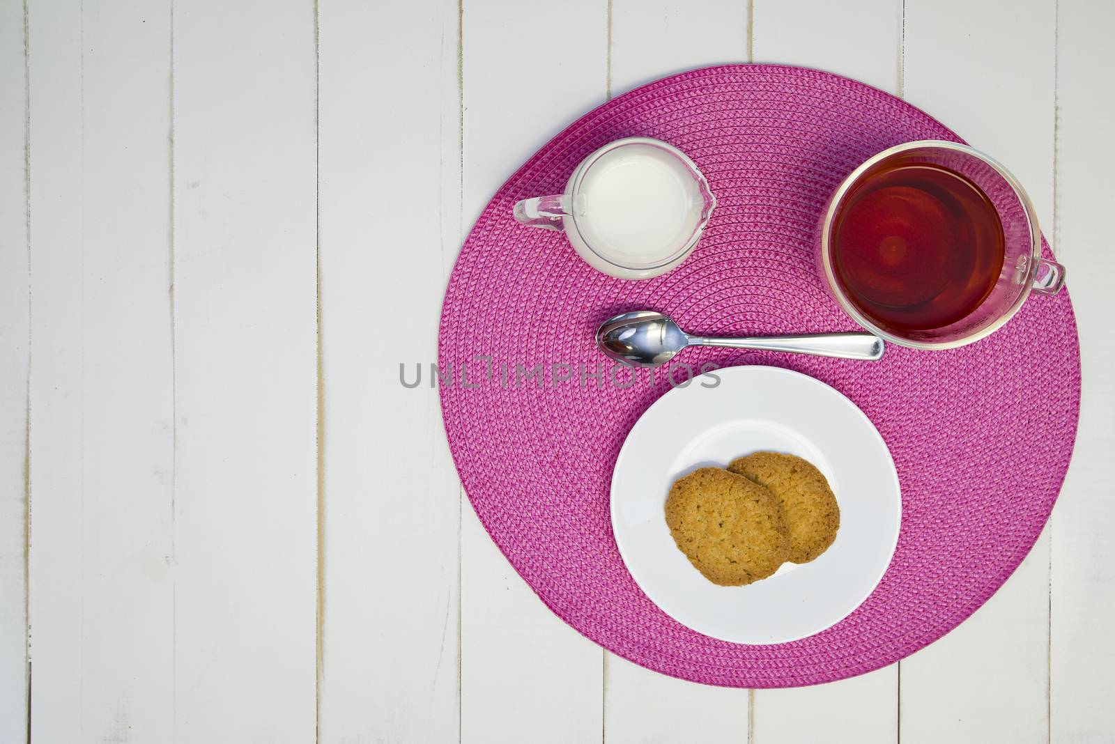 Hot tea, cookies, and milk are arranged on a pink placemat on a white board table. The arrangement is off-center.
