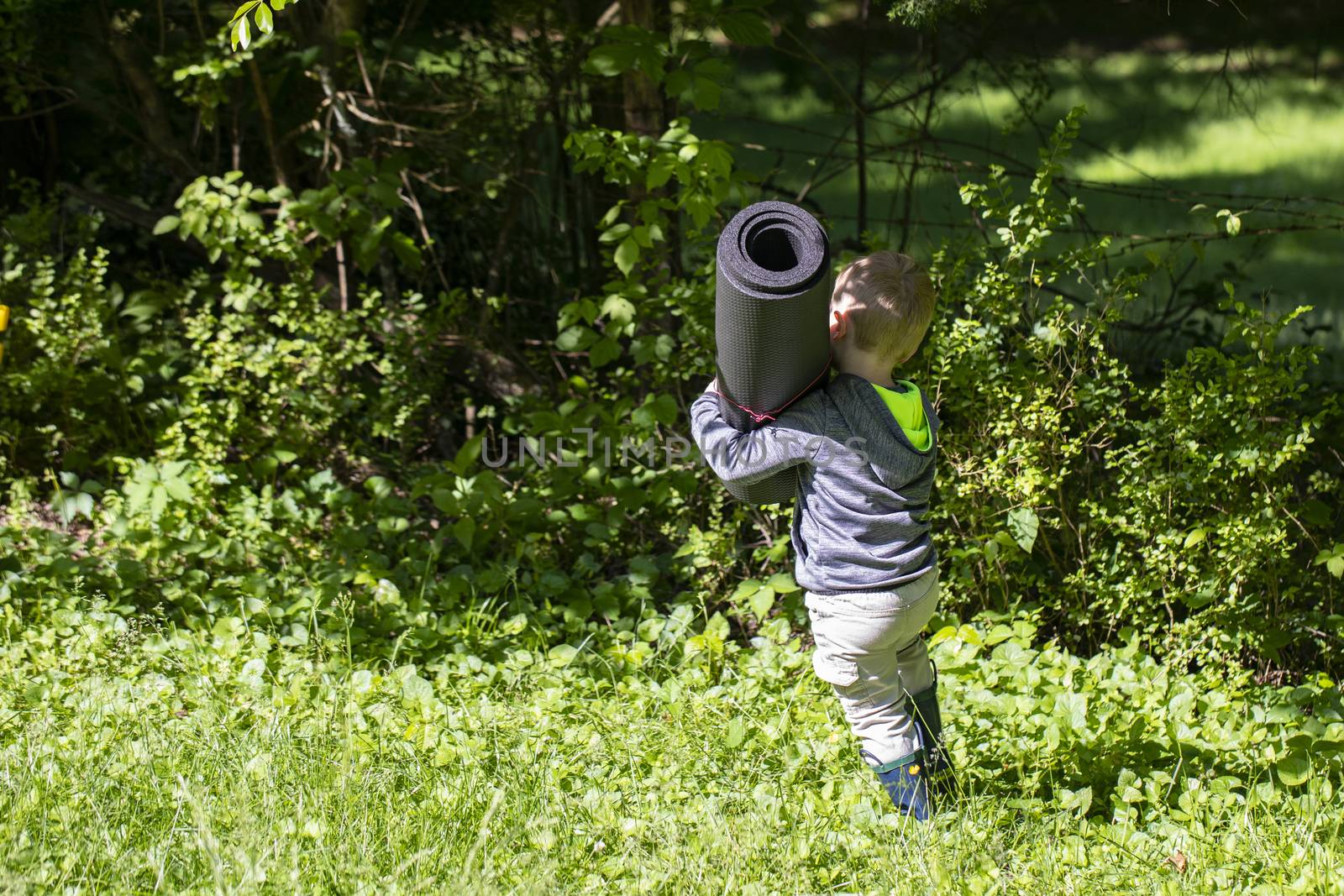 Male toddler carries rolled up mat through the grass.
