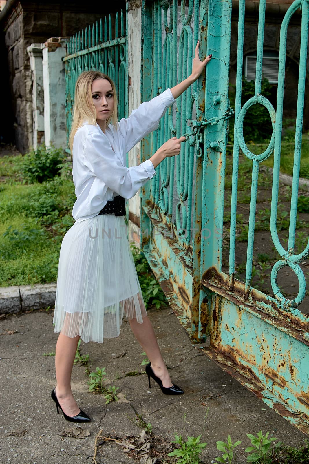 Young blonde woman in white skirt and shirt near the green vintage gate. Young woman's modern portrait.