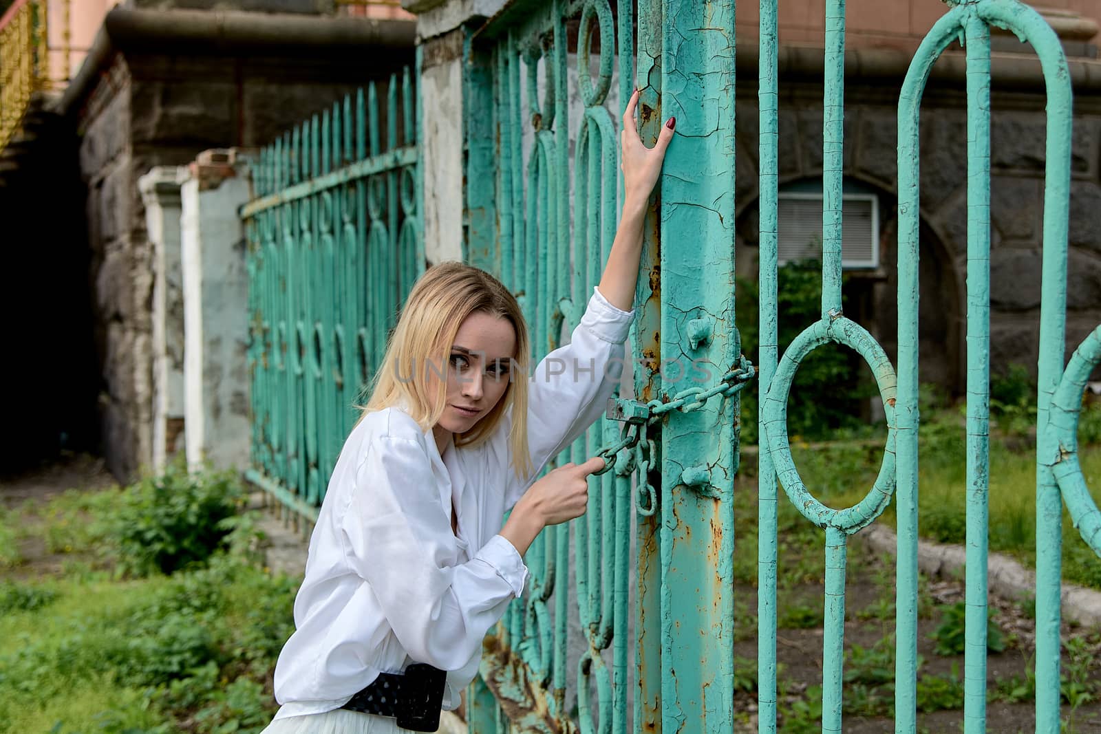 Young blonde woman in white skirt and shirt near the green vintage gate. Young woman's modern portrait.