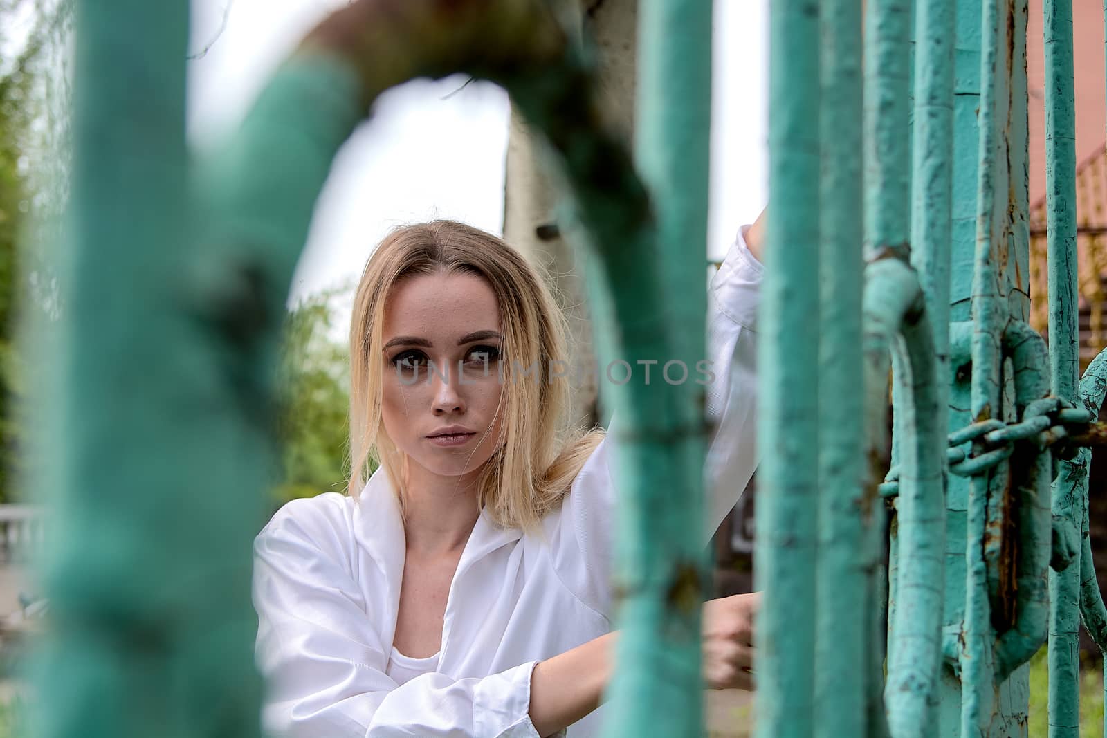 Young blonde woman in white skirt and shirt near the green vintage gate. Young woman's modern portrait.