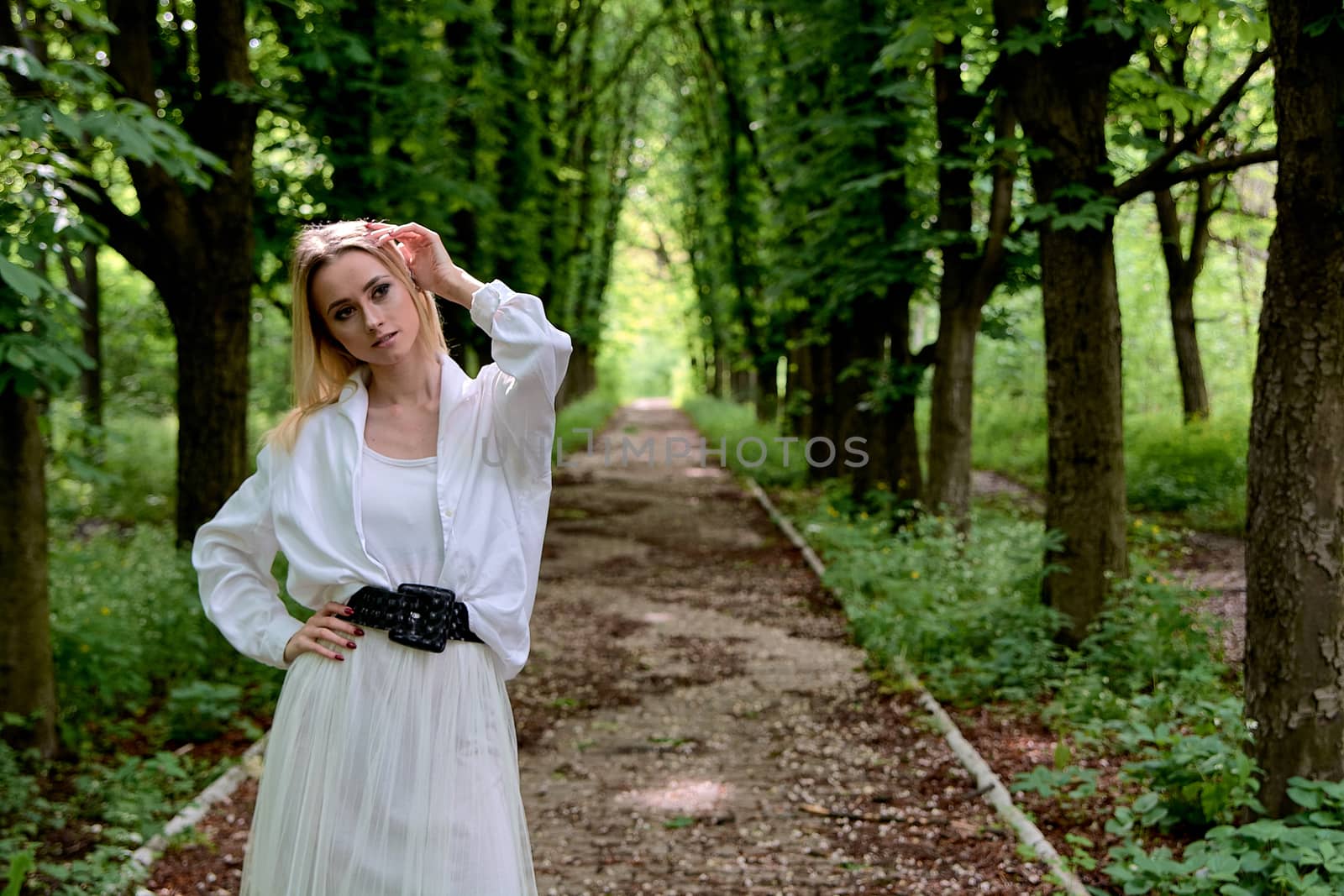 Young blonde woman in white skirt and shirt walks alone along the chestnut alley in the city park. Fashion woman. Young woman's modern portrait.