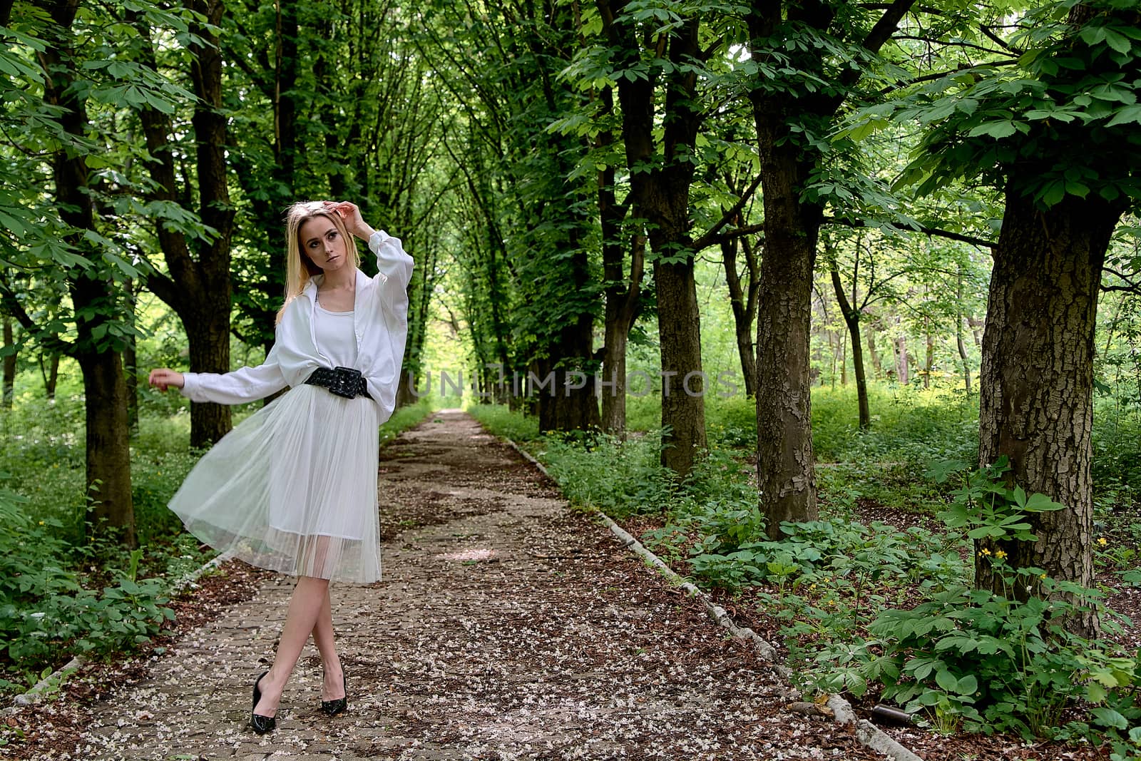 Young blonde woman in white skirt and shirt walks alone along the chestnut alley in the city park. Fashion woman. Young woman's modern portrait.