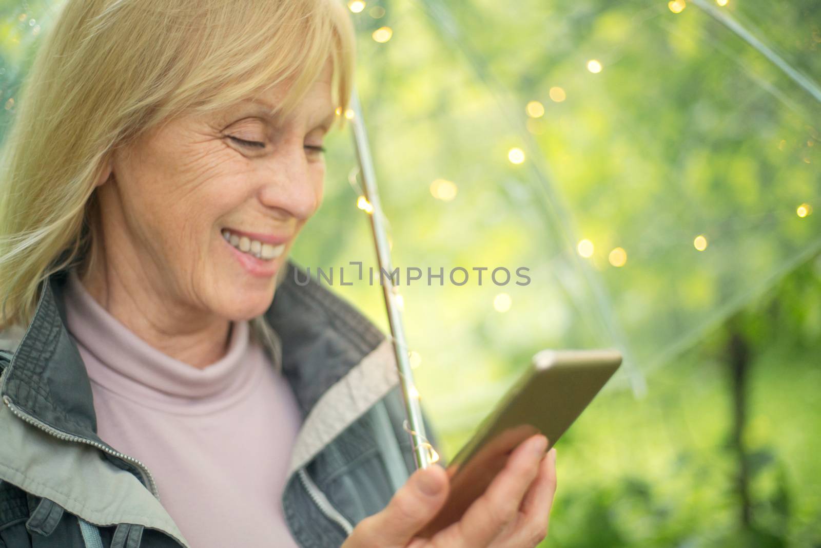 Smiling mature woman reading message on her mobile phone while standing with transparent umbrella with light bokeh background