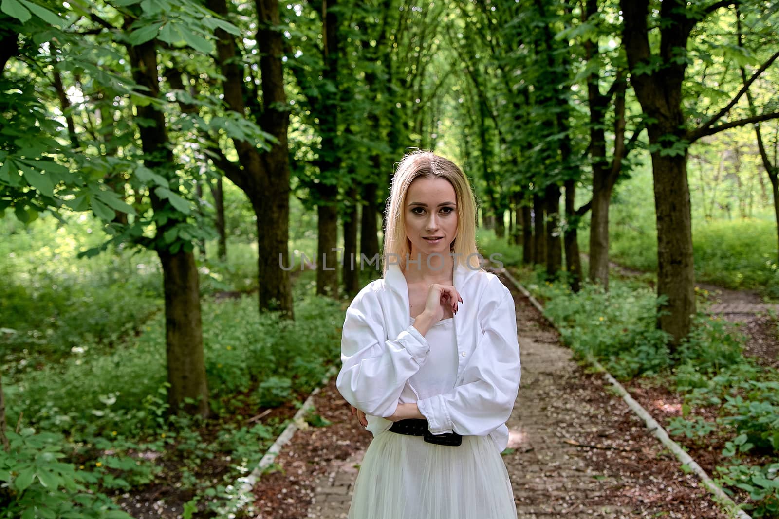 Young blonde woman in white skirt and shirt walks alone along the chestnut alley in the city park. Fashion woman. Young woman's modern portrait.