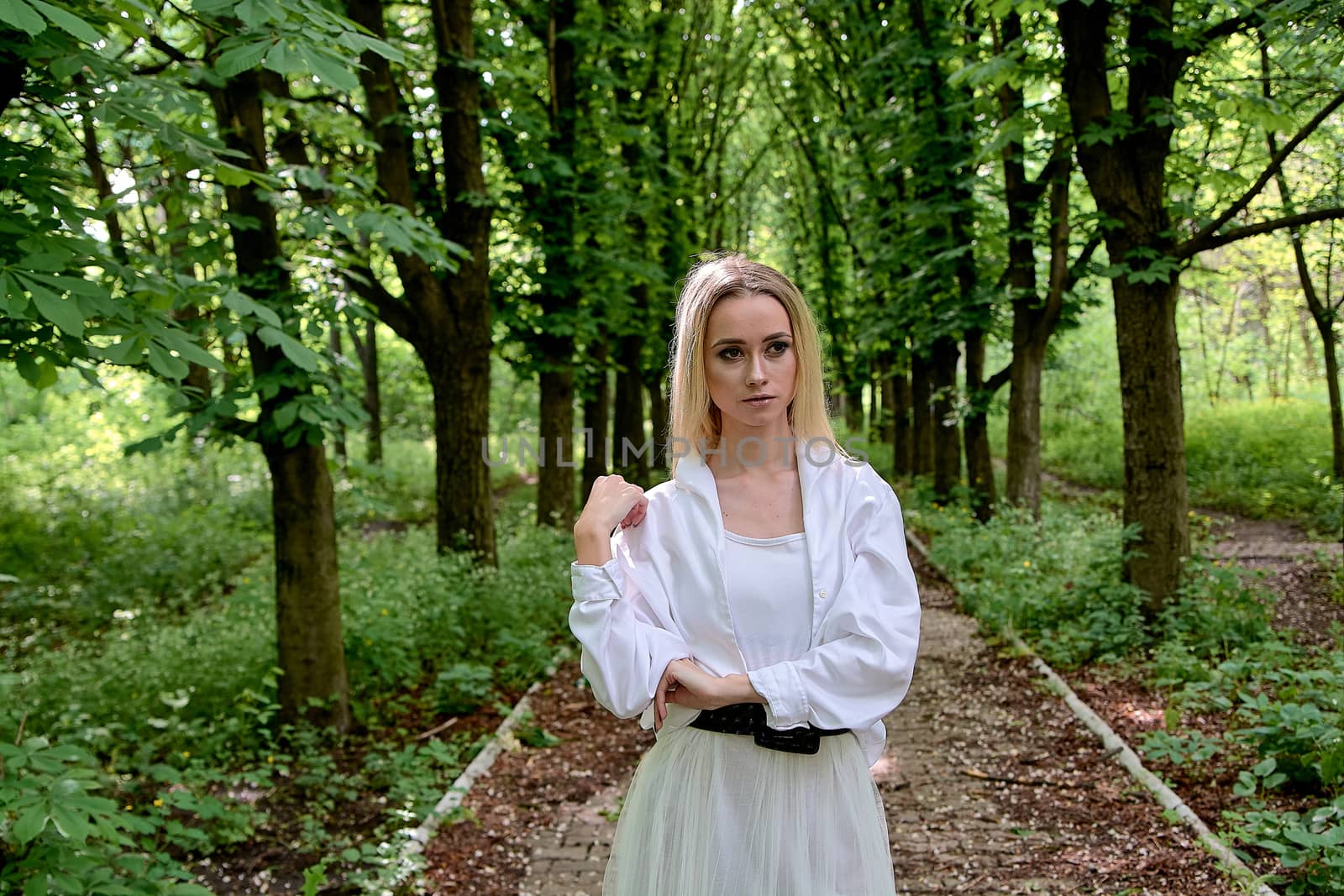 Young blonde woman in white skirt and shirt walks alone along the chestnut alley in the city park. Fashion woman. Young woman's modern portrait.