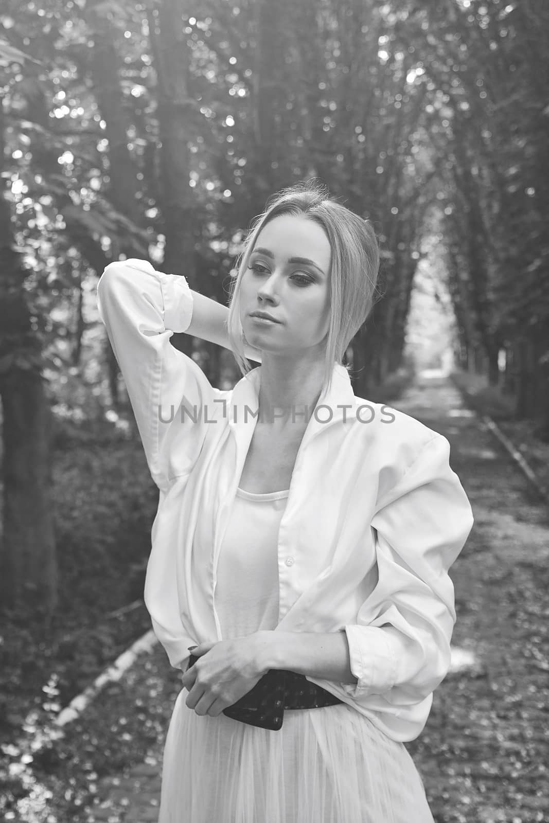 Young blonde woman in white skirt and shirt walks alone along the chestnut alley in the city park. Fashion woman. Young woman's modern portrait.