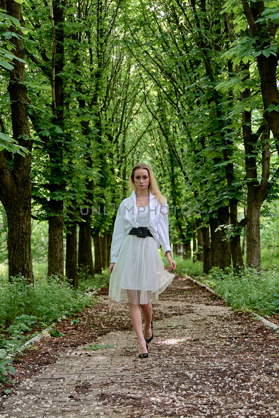 Young blonde woman in white skirt and shirt walks alone along the chestnut alley in the city park. Fashion woman. Young woman's modern portrait.