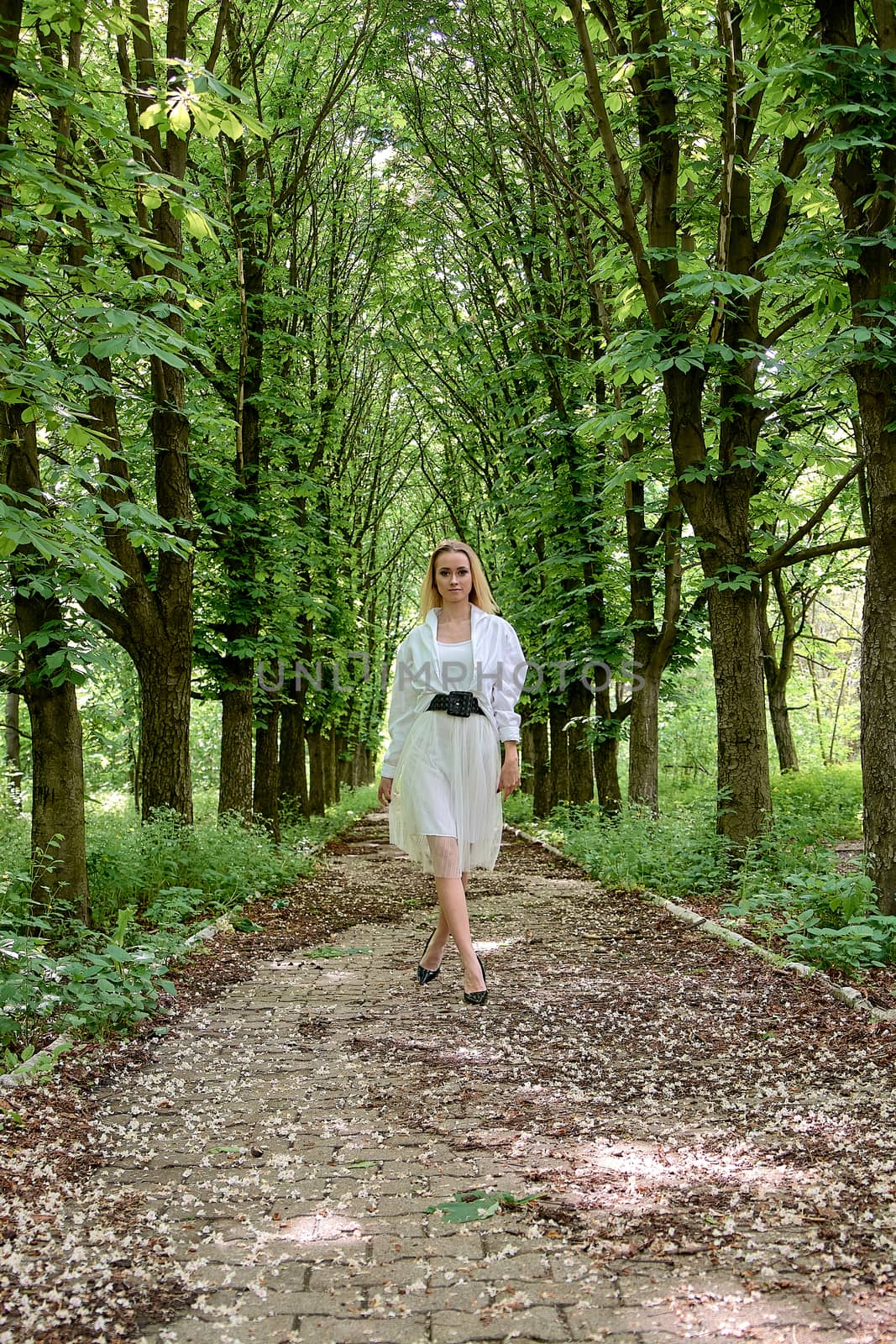 Young blonde woman in white skirt and shirt walks alone along the chestnut alley in the city park. Fashion woman. Young woman's modern portrait.