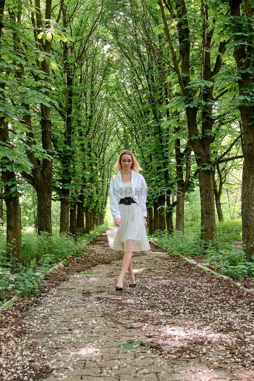 Young blonde woman in white skirt and shirt walks alone along the chestnut alley in the city park. Fashion woman. Young woman's modern portrait.