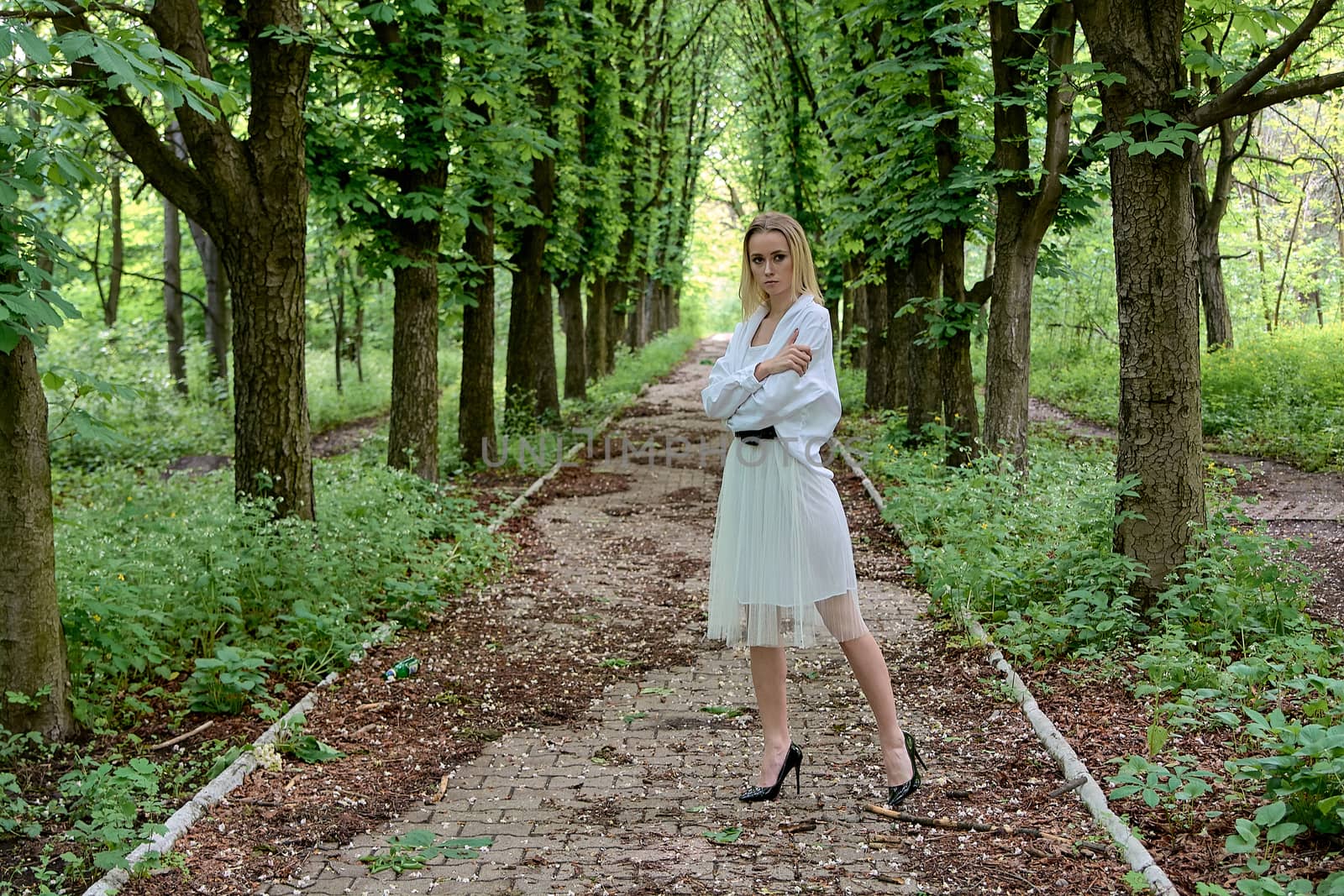 Young blonde woman in white skirt and shirt walks alone along the chestnut alley in the city park. Fashion woman. Young woman's modern portrait.