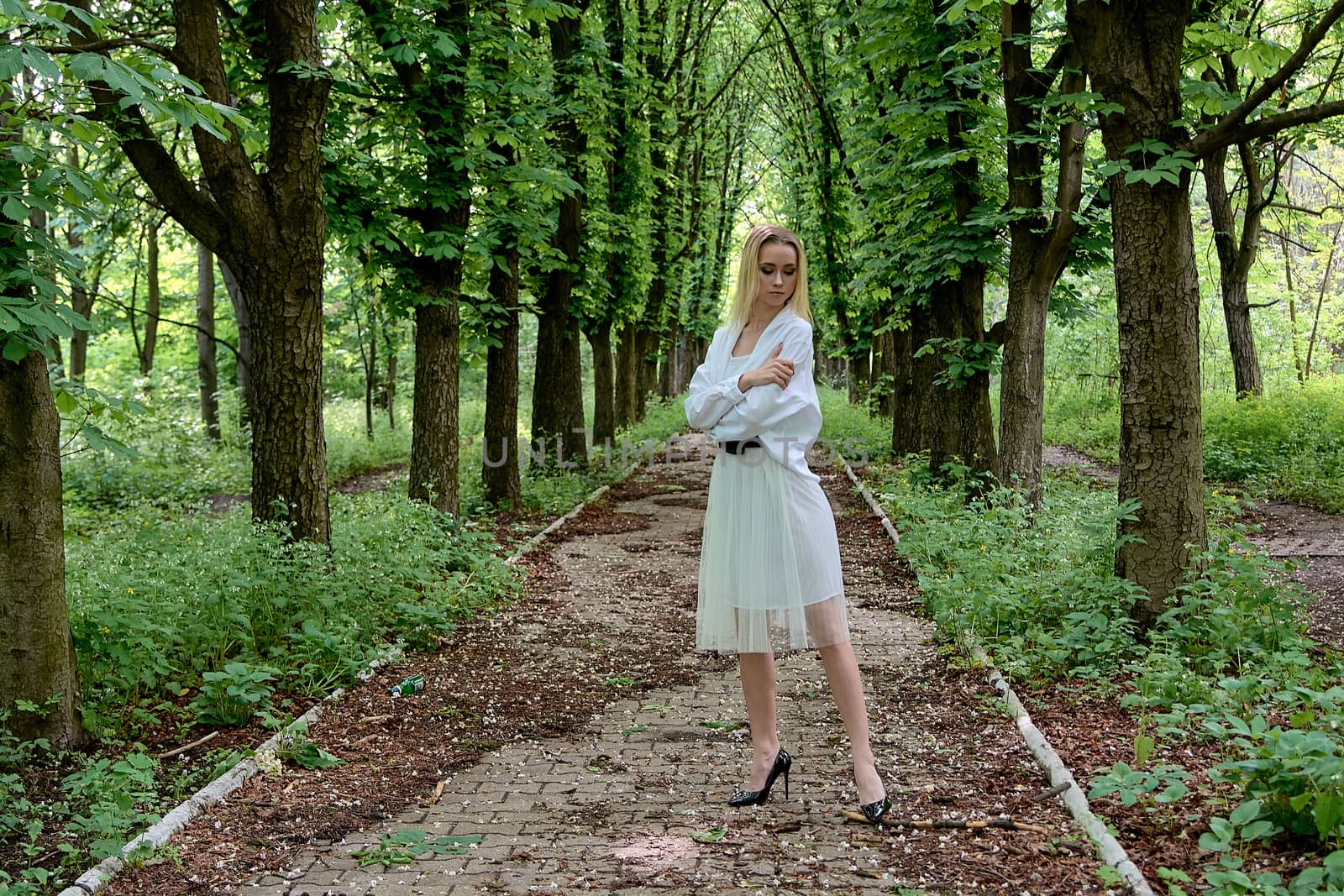 Young blonde woman in white skirt and shirt walks alone along the chestnut alley in the city park. Fashion woman. Young woman's modern portrait.