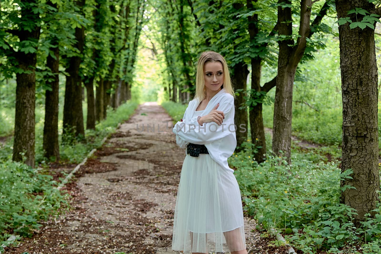 Young blonde woman in white skirt and shirt walks alone along the chestnut alley in the city park. Fashion woman. Young woman's modern portrait.