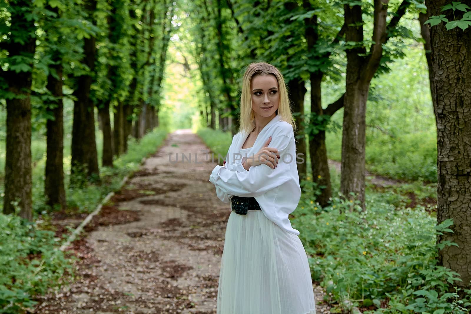 Young blonde woman in white skirt and shirt walks alone along the chestnut alley in the city park. Fashion woman. Young woman's modern portrait.