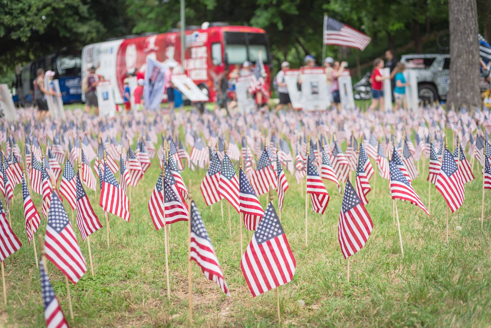 Close-up view of lawn American flag with row of people from Memorial Day March event in Dallas, Texas, USA. Blurry crowded family members carry fallen heroes banners pictures placards in parade