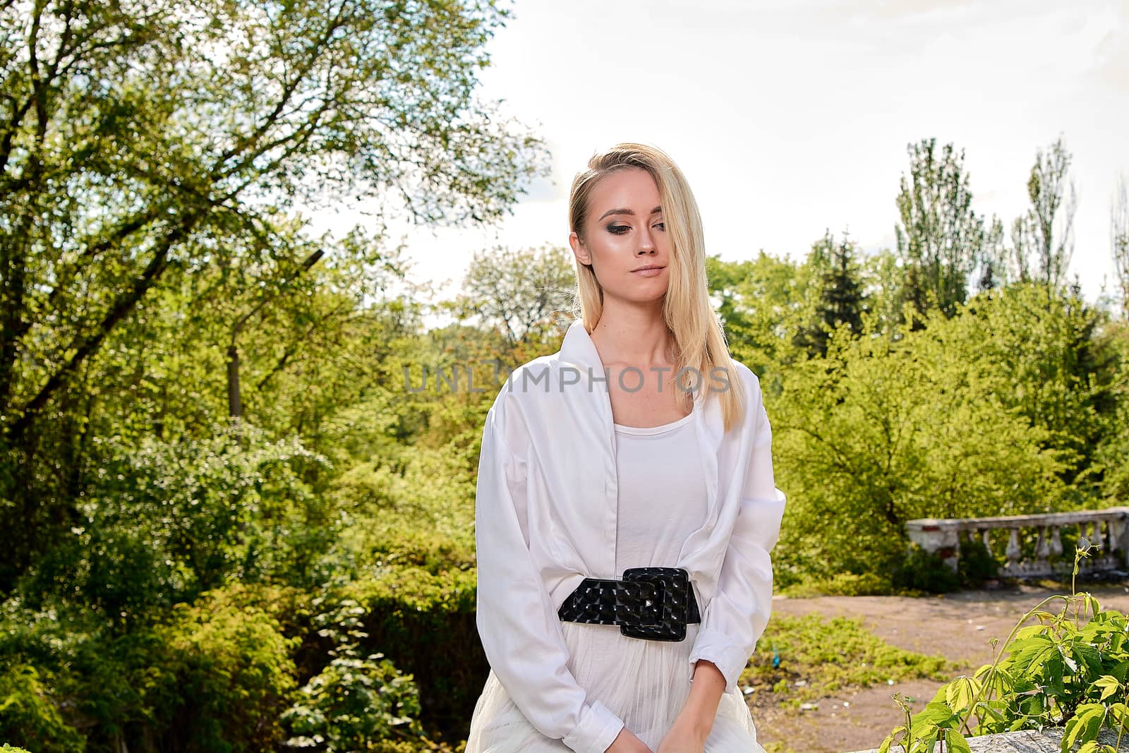 Young blonde woman in white skirt and shirt in the old sunny summer city park. Fashion woman. Young woman's modern portrait.