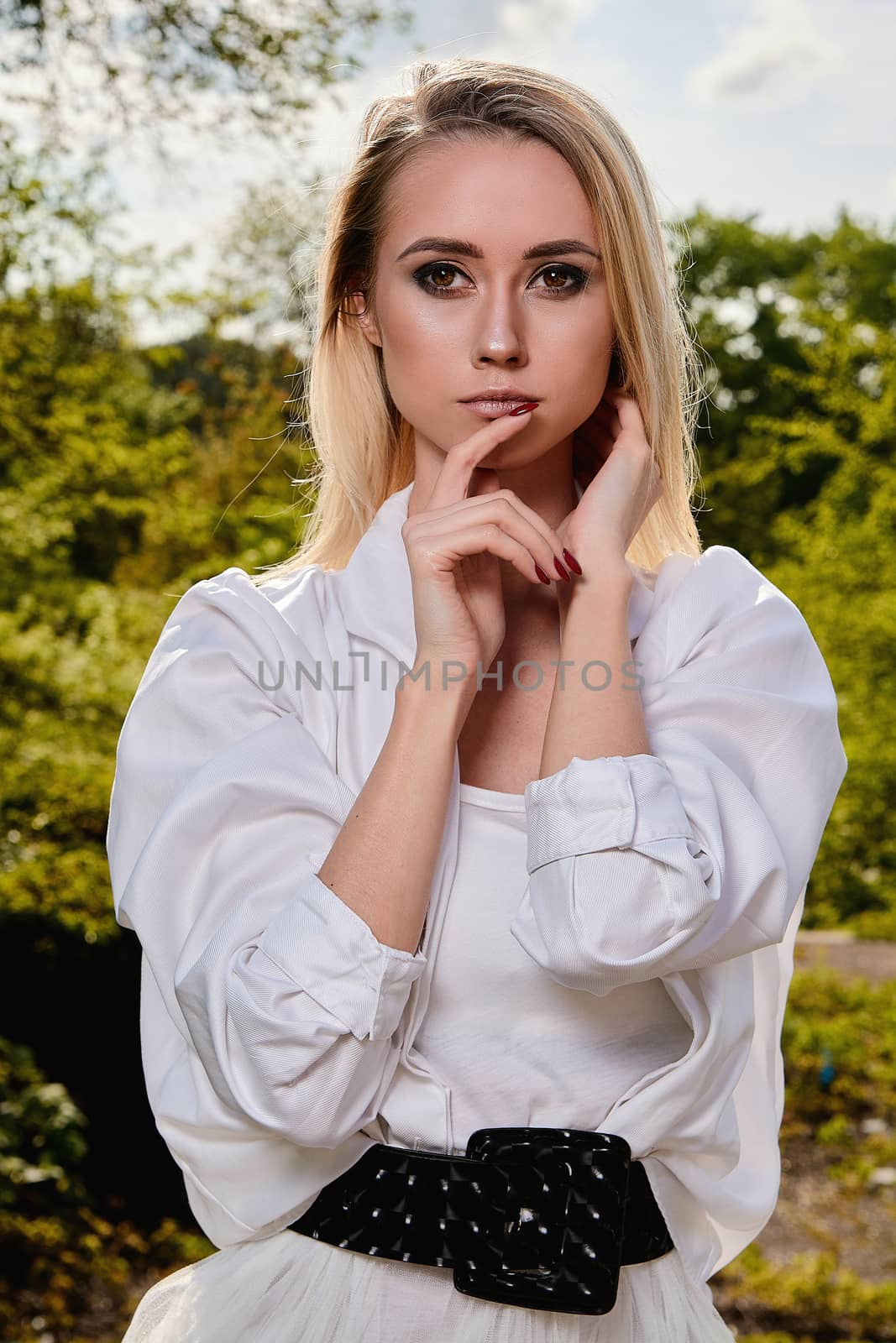 Young blonde woman in white skirt and shirt in the old sunny summer city park. Fashion woman. Young woman's modern portrait.