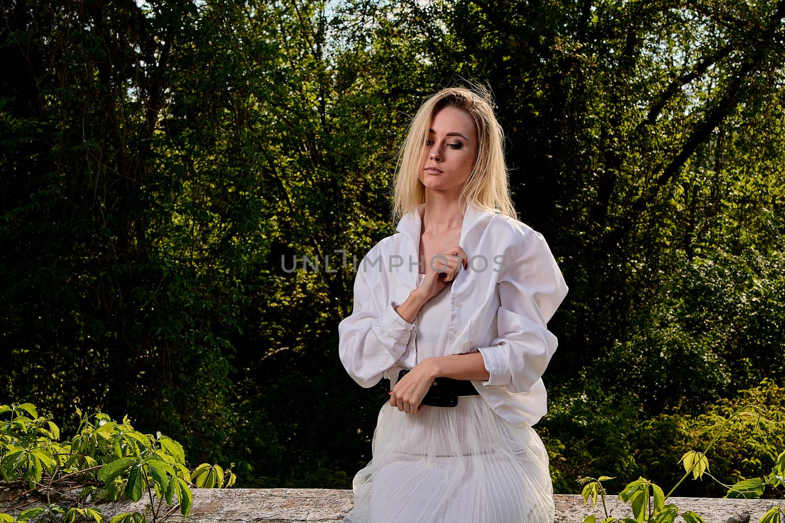 Young blonde woman in white skirt and shirt in the old sunny summer city park. Fashion woman. Young woman's modern portrait.