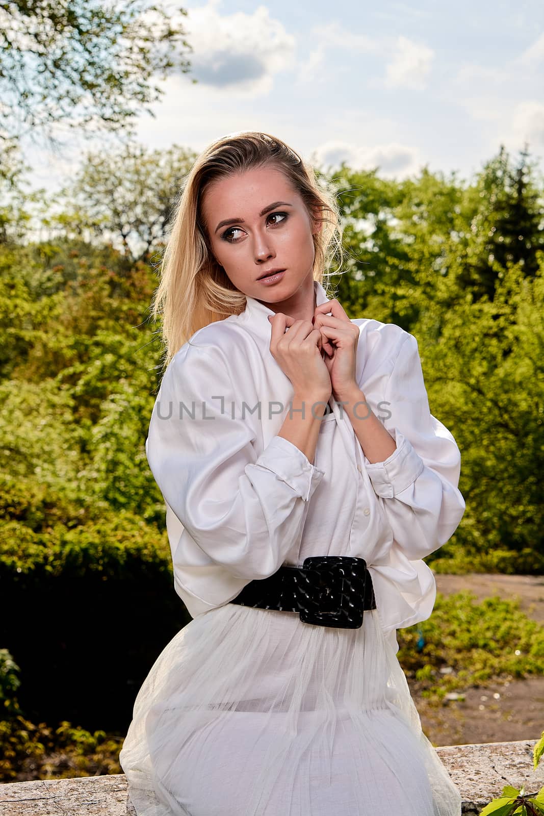 Young blonde woman in white skirt and shirt in the old sunny summer city park. Fashion woman. Young woman's modern portrait.