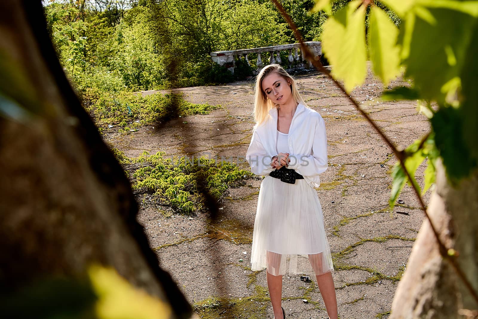 Young blonde woman in white skirt and shirt in the old sunny summer city park. Fashion woman. Young woman's modern portrait.