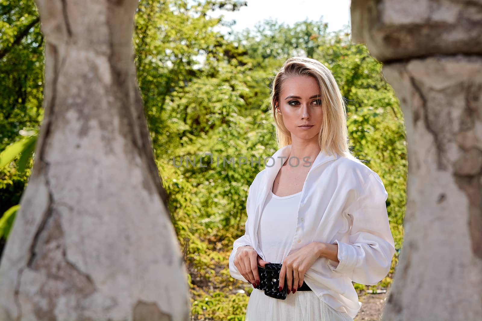 Young blonde woman in white skirt and shirt in the old sunny summer city park. Fashion woman. Young woman's modern portrait.