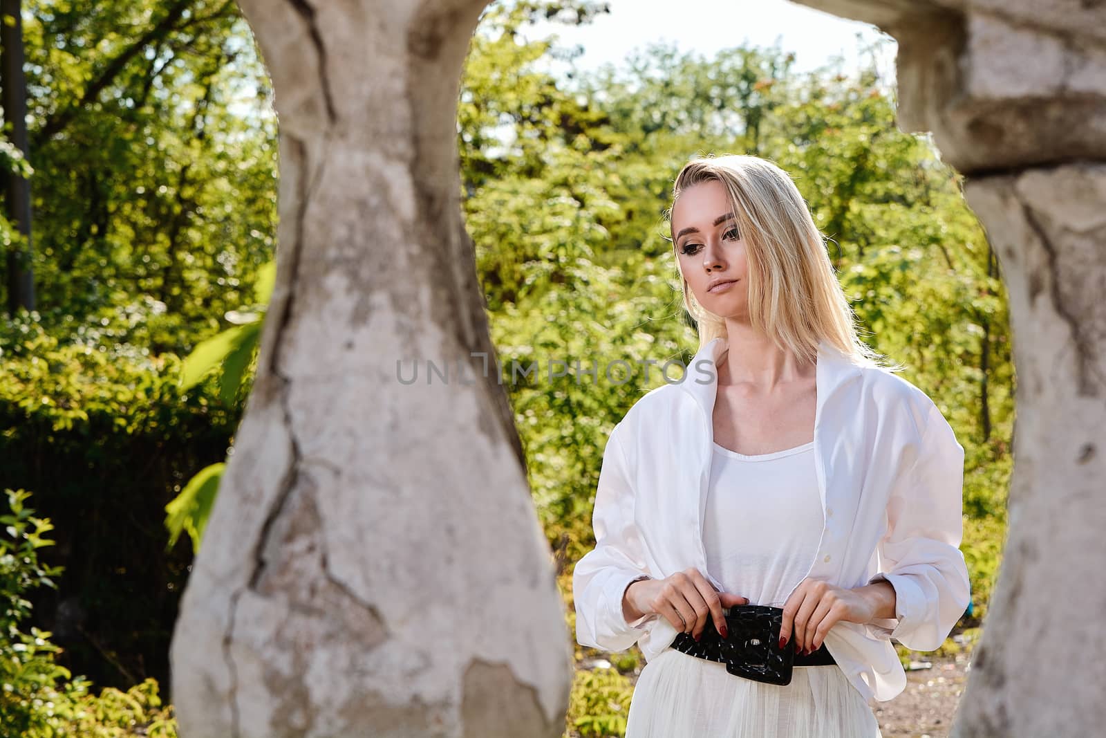 Young blonde woman in white skirt and shirt in the old sunny summer city park. Fashion woman. Young woman's modern portrait.