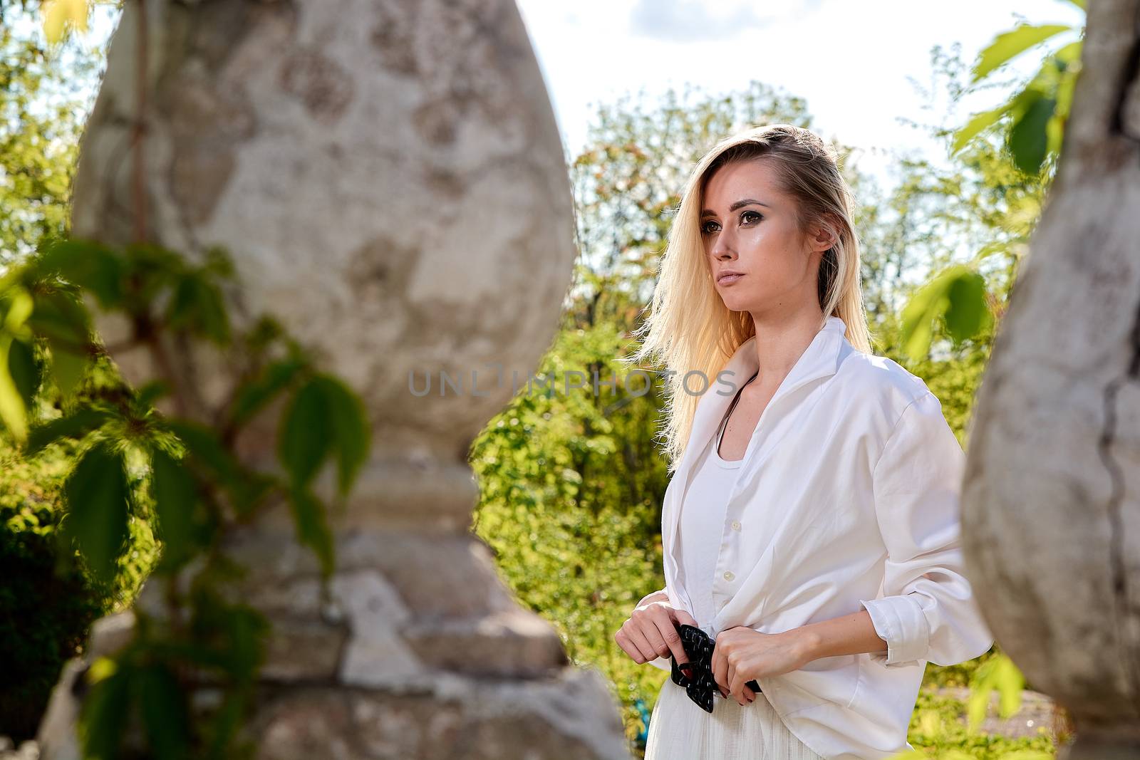 Young blonde woman in white skirt and shirt in the old sunny summer city park. Fashion woman. Young woman's modern portrait.