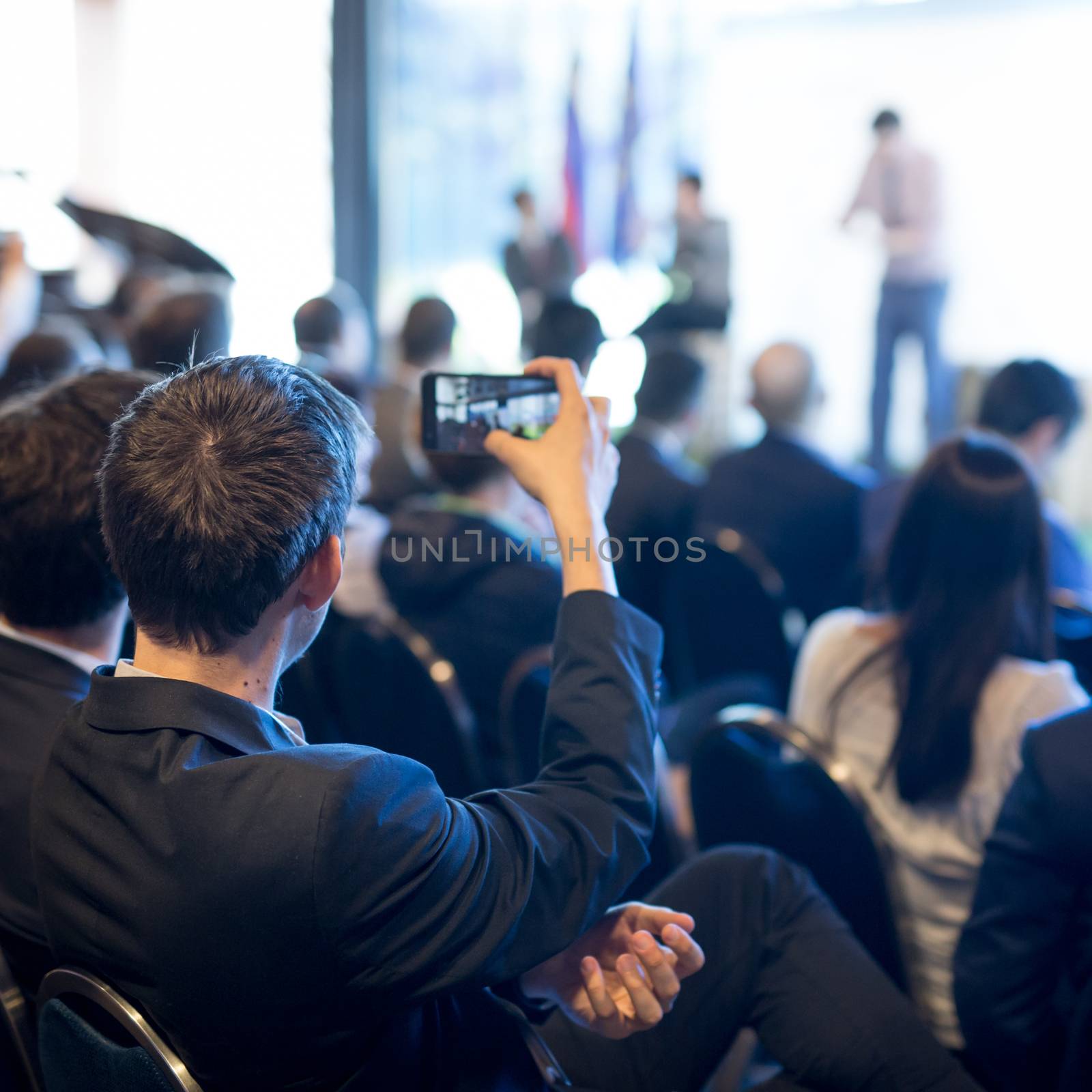Businessman takes a picture of corporate business presentation at conference hall using smartphone. Business and Entrepreneurship concept. Focus on unrecognizable person in audience.