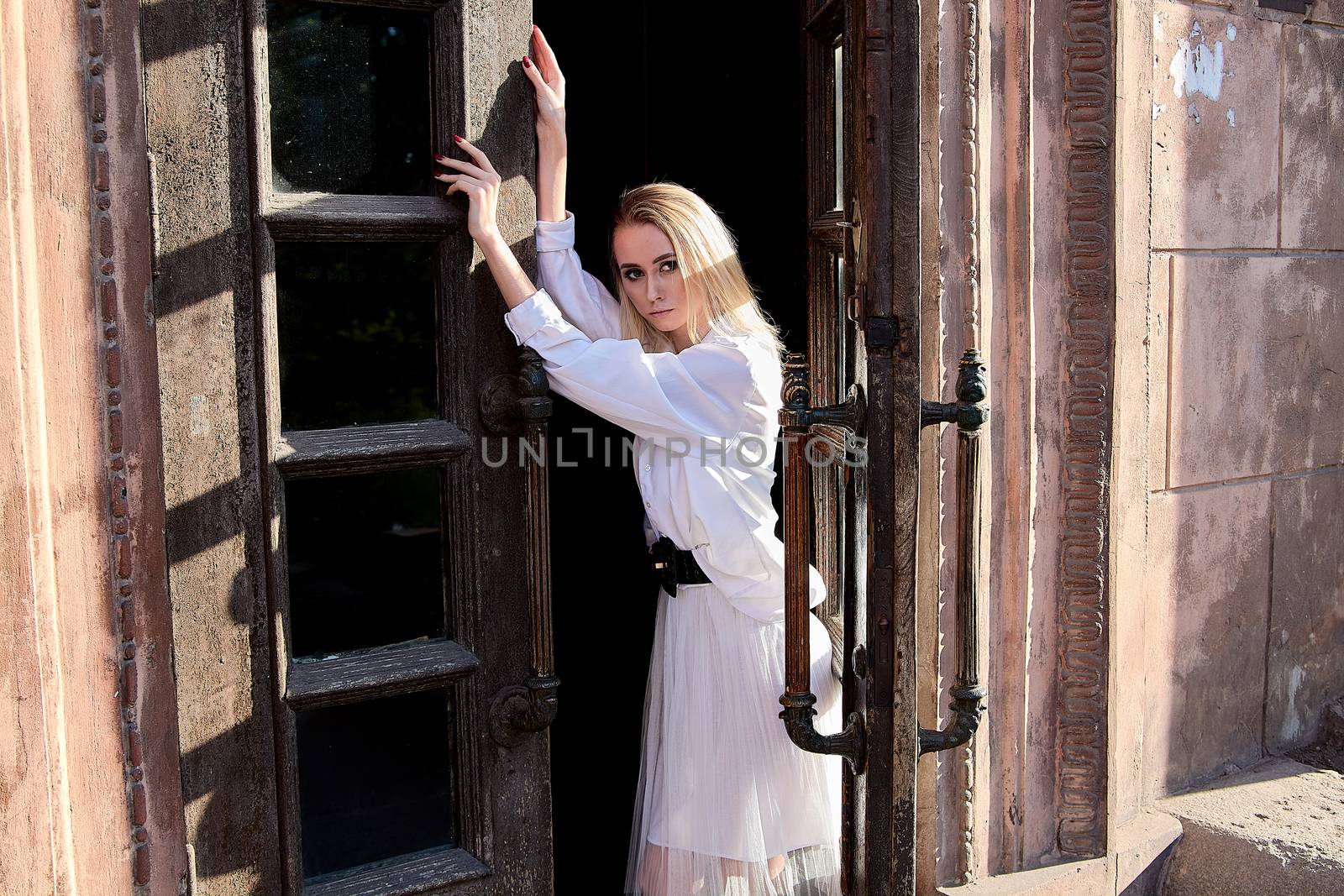 Young blonde woman in white skirt and shirt stands in the old wooden doorway. The old wooden door. Fashion woman. Young woman modern portrait.