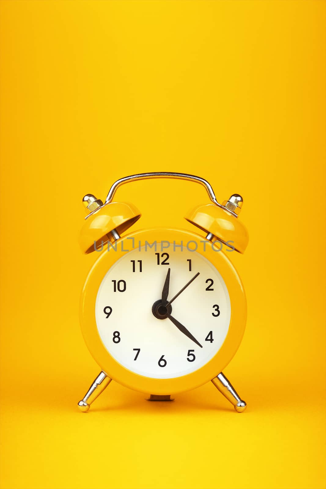 Close up one small yellow metal twin bell retro alarm clock over warm yellow paper background with copy space, low angle front view