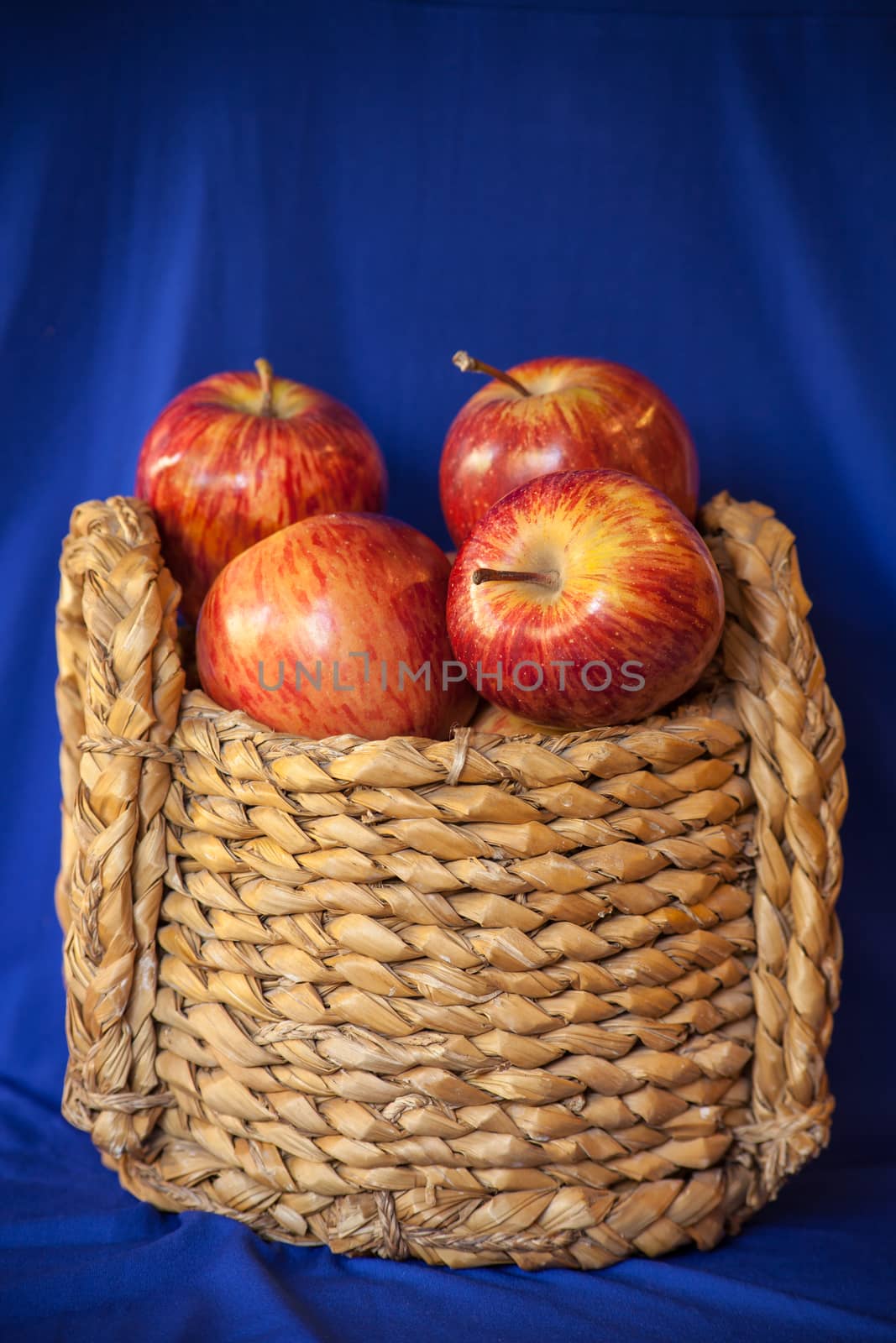 Still life image of a small grass basket filled with large red Starking apples on a blue background.