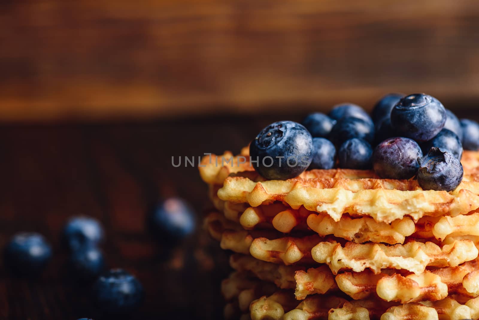 Blueberries on the Top of the Waffles Stack and Other Scattered on Wooden Background. Copy Space on the Left.