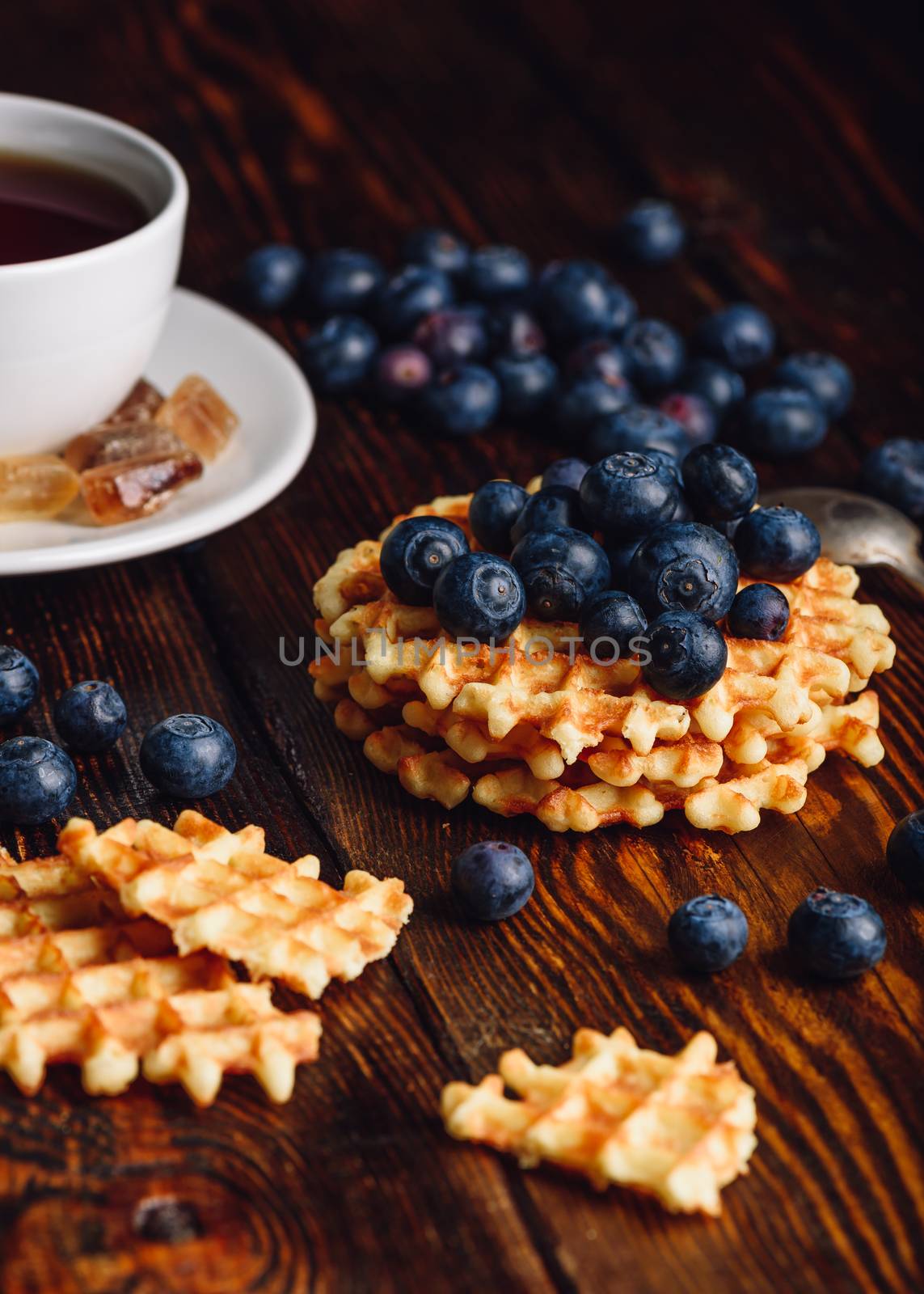 Cup of Tea with Blueberries on the Top of the Waffles Stack. Other Berries and Broken Waffle Scattered on Wooden Background.