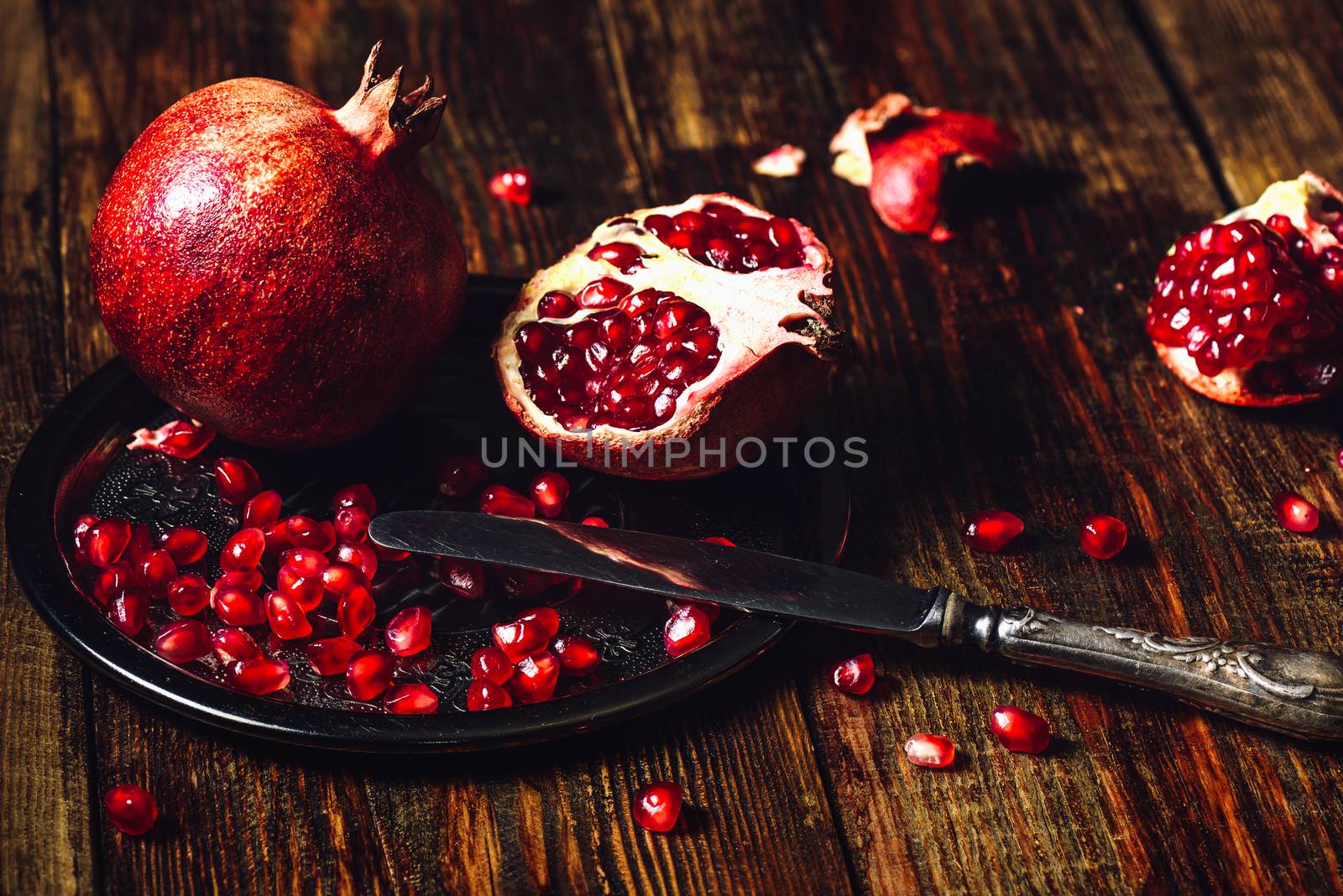 Opened Pomegranate and Whole One wiith Seeds on Metal Plate and Vintage Knife. Some Seeds and Pieces Scattered on Wooden Surface.
