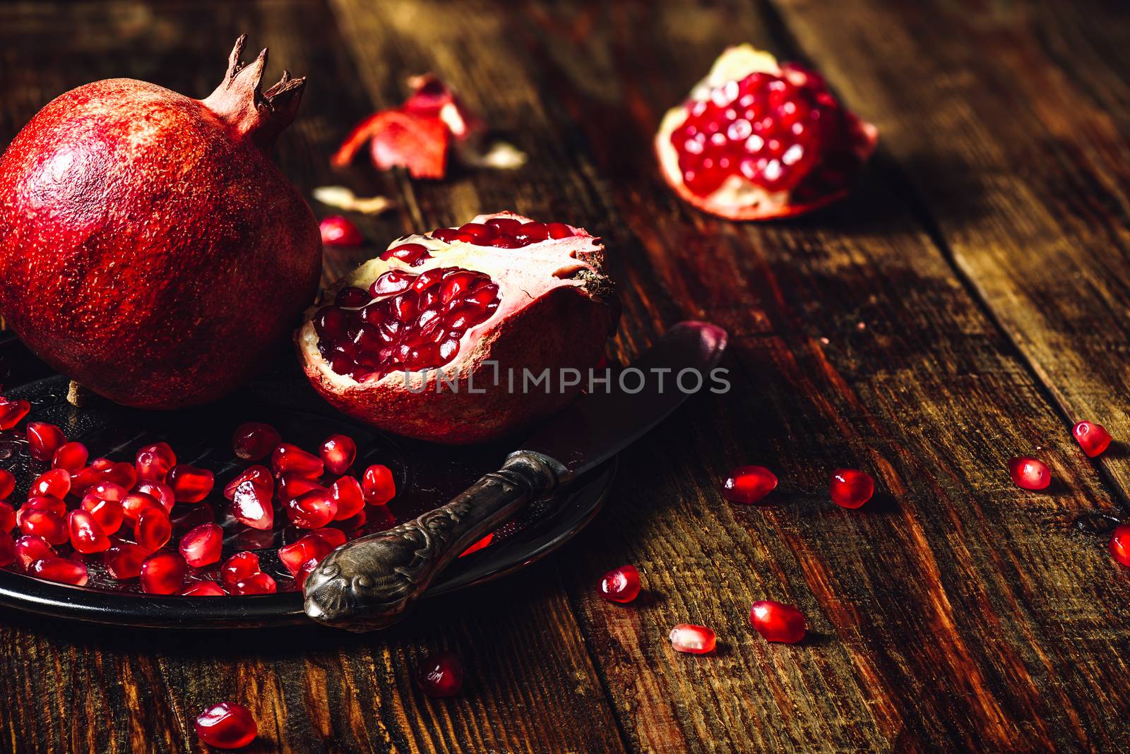 Opened Pomegranate and Whole One wiith Seeds on Metal Plate and Vintage Knife. Some Seeds and Pieces Scattered on Wooden Surface. Copy Space on the Right.