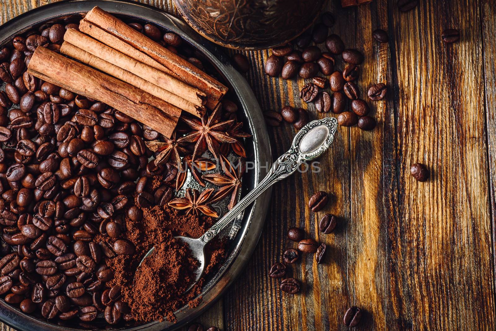 Coffee Beans with Spooonful of Ground Coffee, Cinnamon Sticks and Chinese Star Anise on Metal Plate. Some Beans Scattered on Wooden Table. View from Above.