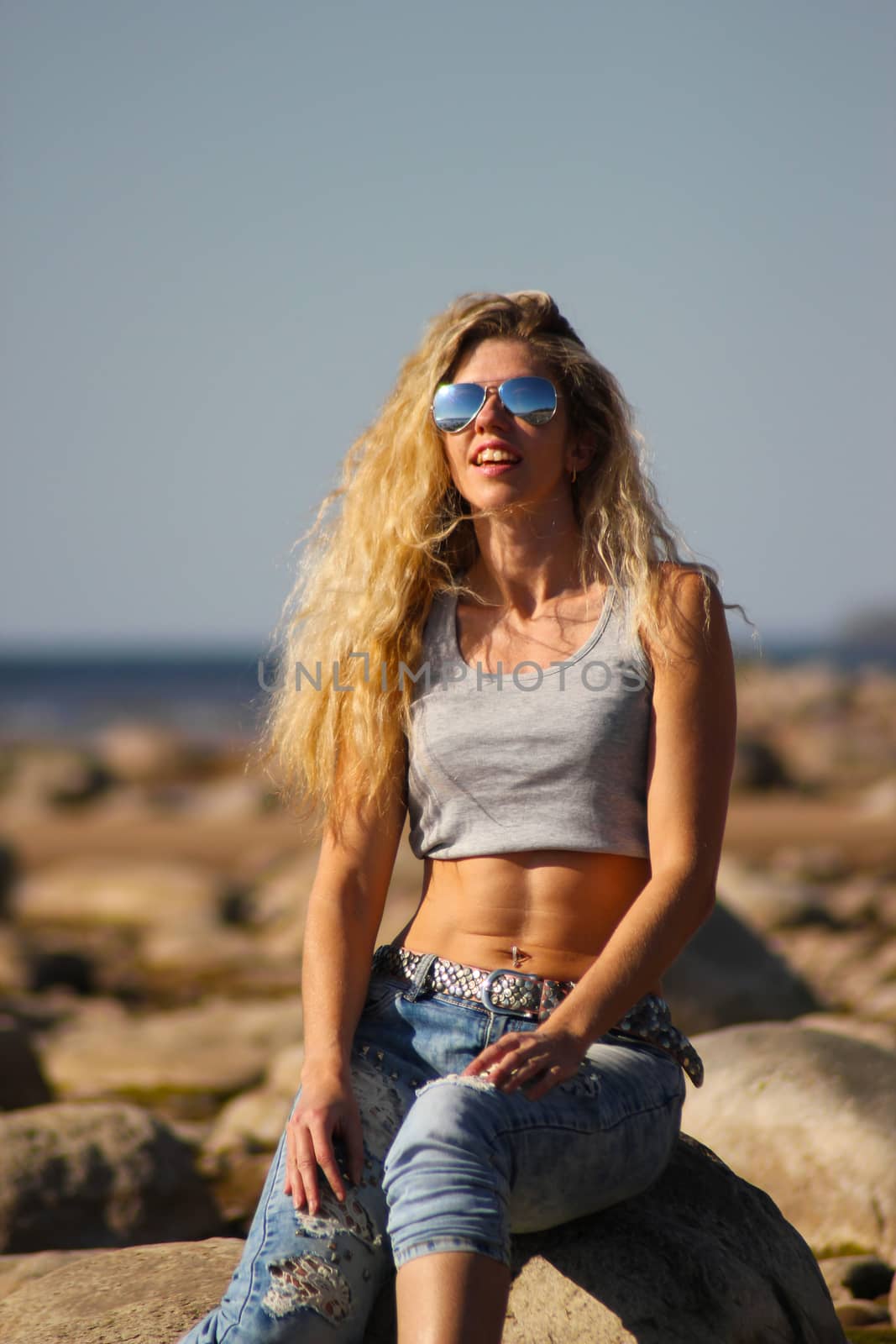 Tanned blonde girl in sunglasses and jeans sits on the rocks on the beach on a sunny day. by zakob337