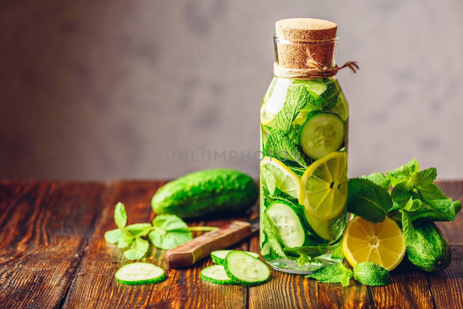 Bottle of Infusion with Sliced Lemon, Cucumber and Fresh Mint Leaves. Ingredients and Knife on Table. Copy Space on the Left.