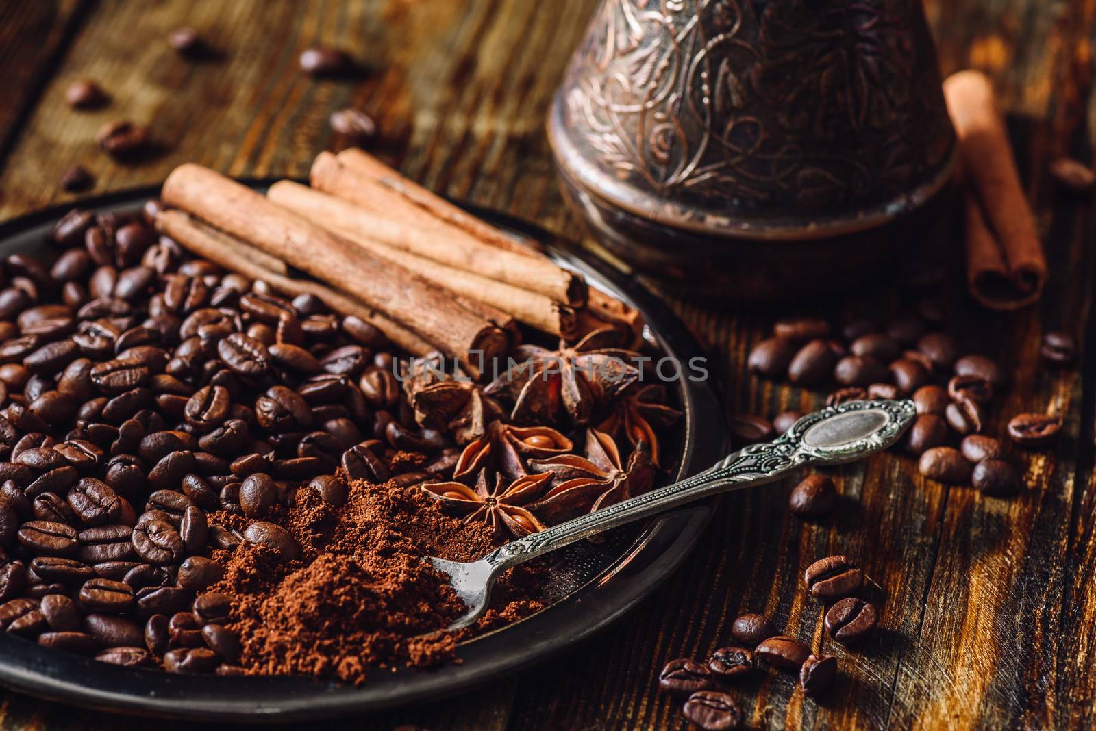 Coffee Beans with Spooonful of Ground Coffee, Cinnamon Sticks and Chinese Star Anise on Metal Plate. Beans Scattered on Wooden Table and Cezve on Backdrop.