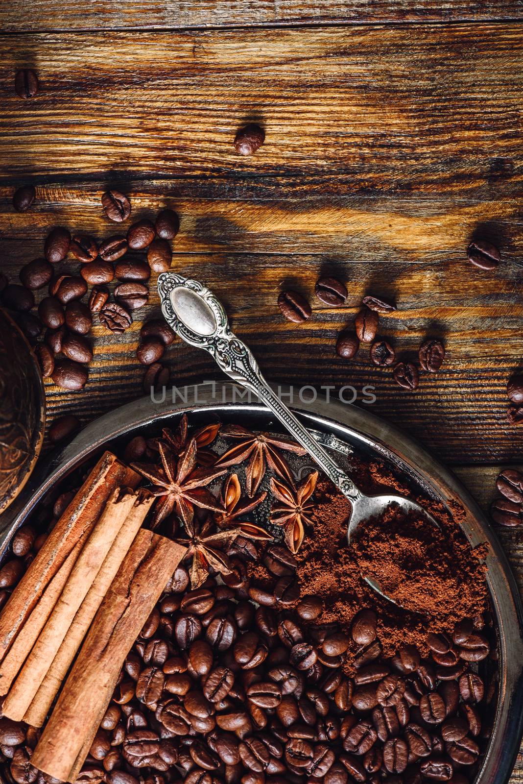 Coffee Beans with Spooonful of Ground Coffee, Cinnamon Sticks and Star Anise on Plate. Some Beans Scattered on Wooden Table. View from Above. Vertical Orientation.